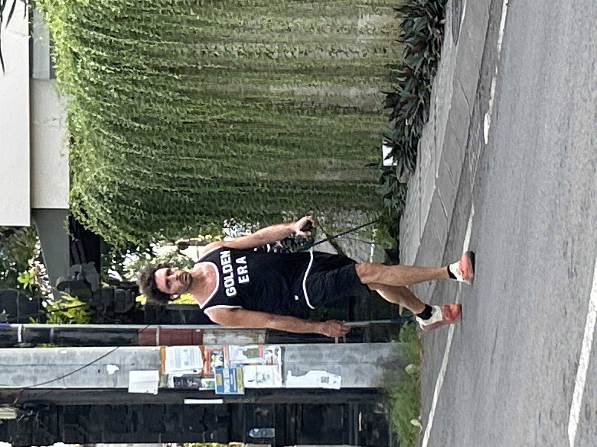 A man with curly dark hair and a beard is jogging on a paved road, holding a black leash in his right hand. He is wearing a black tank top with white trim and the text "GOLDEN ERA" printed in white on the chest. He is also wearing black athletic shorts with white trim and pink and white running shoes. The man is positioned at an angle, appearing to be in motion.
Behind the man, a lush green wall of hanging plants covers a vertical surface. To his right, the road continues, marked with white lines. On the left side of the image, there is a gray metal structure with some posters or papers attached to it, and glimpses of greenery. The lighting suggests it is daytime, possibly late afternoon given the shadows. The overall scene appears to be outdoors in a tropical or subtropical environment, consistent with the provided location context of Mengwi, Indonesia. The man's posture and expression suggest he is focused on his exercise.
