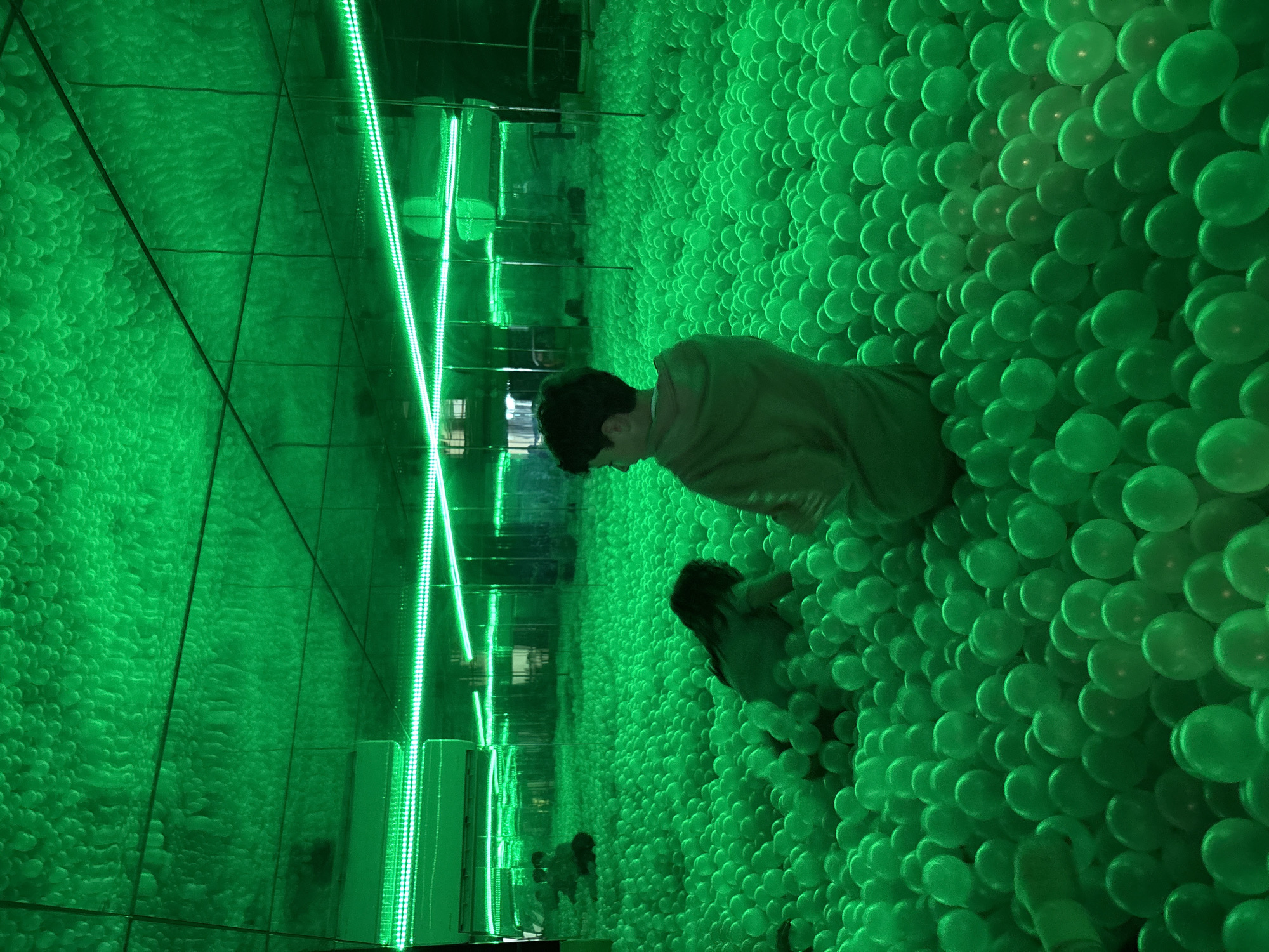 The image shows a room in Mexico City, Mexico filled with green spheres. A person is lying down, partially submerged in the spheres. The room is illuminated with bright green neon lights. The walls and ceiling appear to be mirrored, creating a sense of endless spheres and reflections.