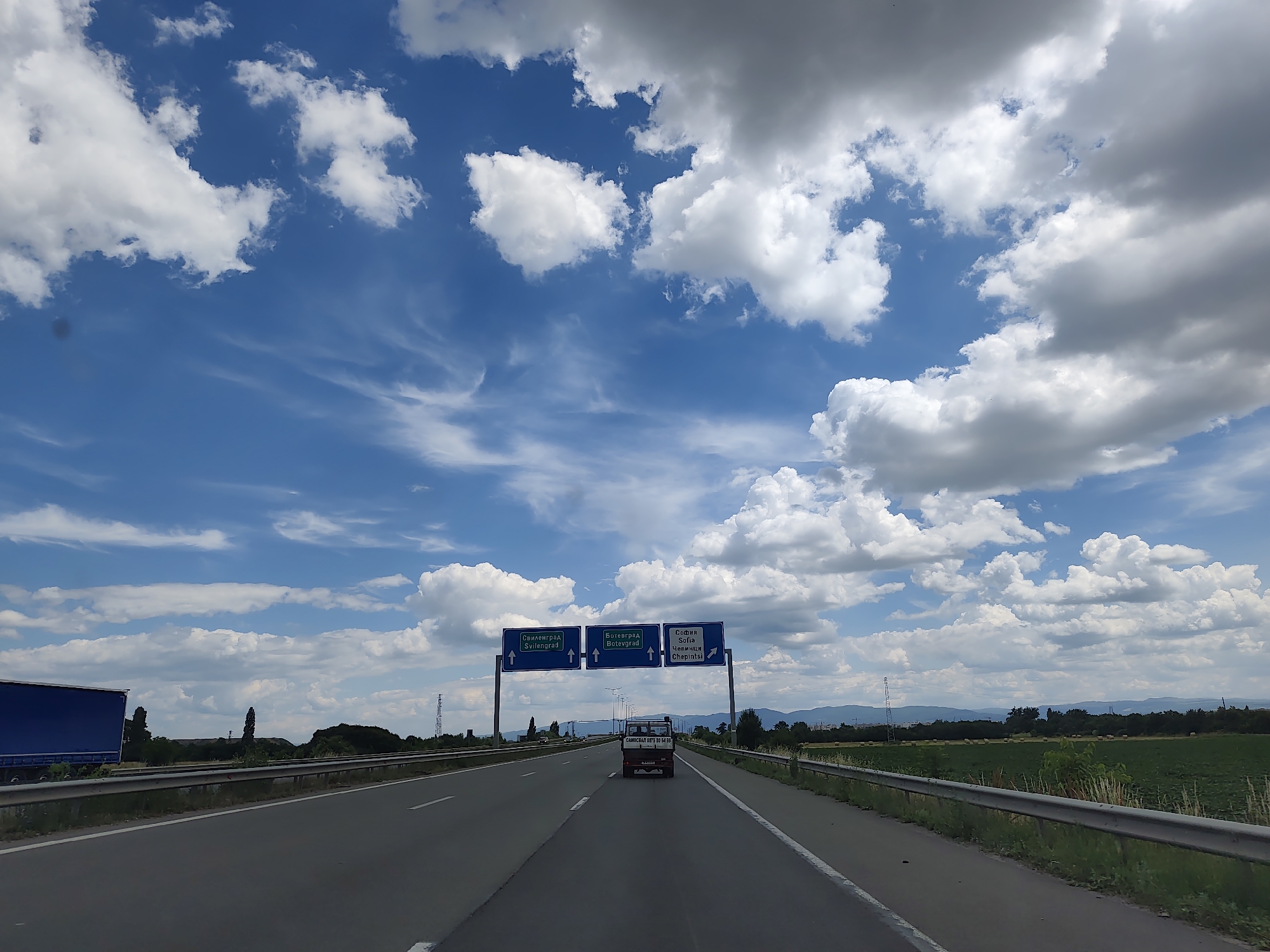 A wide-angle, eye-level, outdoor, daytime shot shows a highway with a bright blue sky filled with white and gray cumulus clouds. The highway is in the foreground, with two lanes visible in each direction, separated by a solid white line. A silver metal guardrail runs along the right side of the highway. In the distance, overhead signs indicate directions to "Svilengrad," "Botevgrad," and "Sofia" (with "Chepinsko" also visible on the Sofia sign). A small, white truck with a dark roof is in the center lane ahead, and a large blue semi-trailer truck is in the left lane on the far left of the frame. The landscape beyond the highway includes green fields, trees, and distant hills or mountains. A power line tower is visible on the right side of the road. The overall scene suggests a journey or travel on a highway, likely near Sofia, Bulgaria, given the road signs.