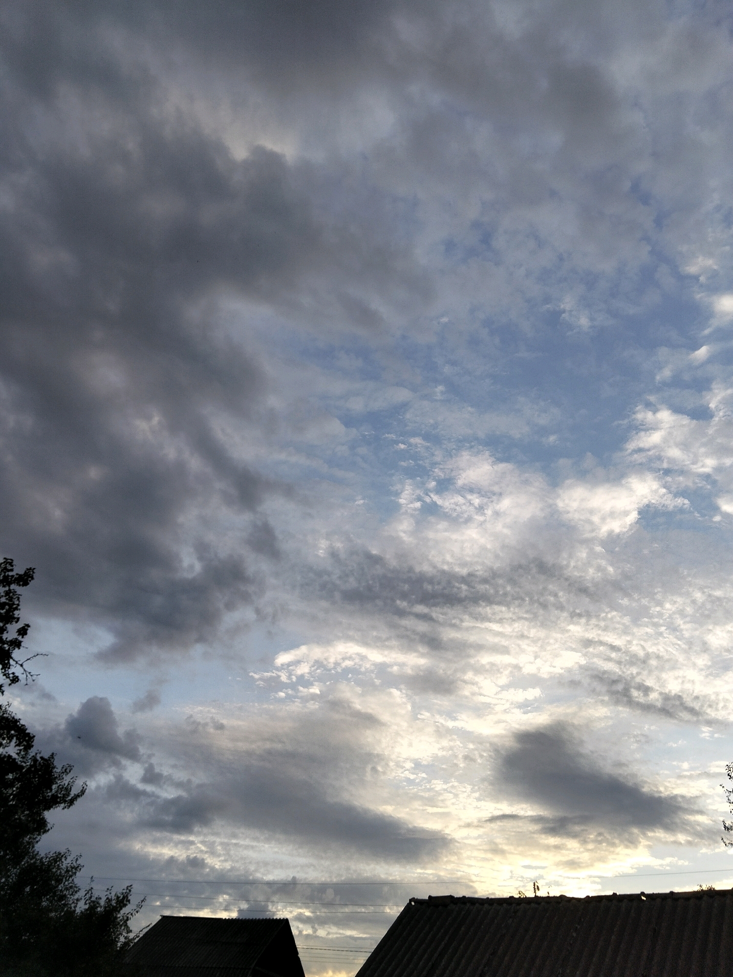 The image captures a dramatic sky over Ustyvytsia, Ukraine, with a mix of dark, heavy clouds on the left and lighter, wispy clouds scattered across a blue sky on the right. The sun appears to be setting or rising, casting a warm glow through the clouds on the right side of the frame. In the foreground, the dark silhouettes of two rooftops are visible, suggesting a residential area. A dark silhouette of a tree with sparse foliage is present on the far left. The scene conveys a sense of impending weather change or the end of a day.