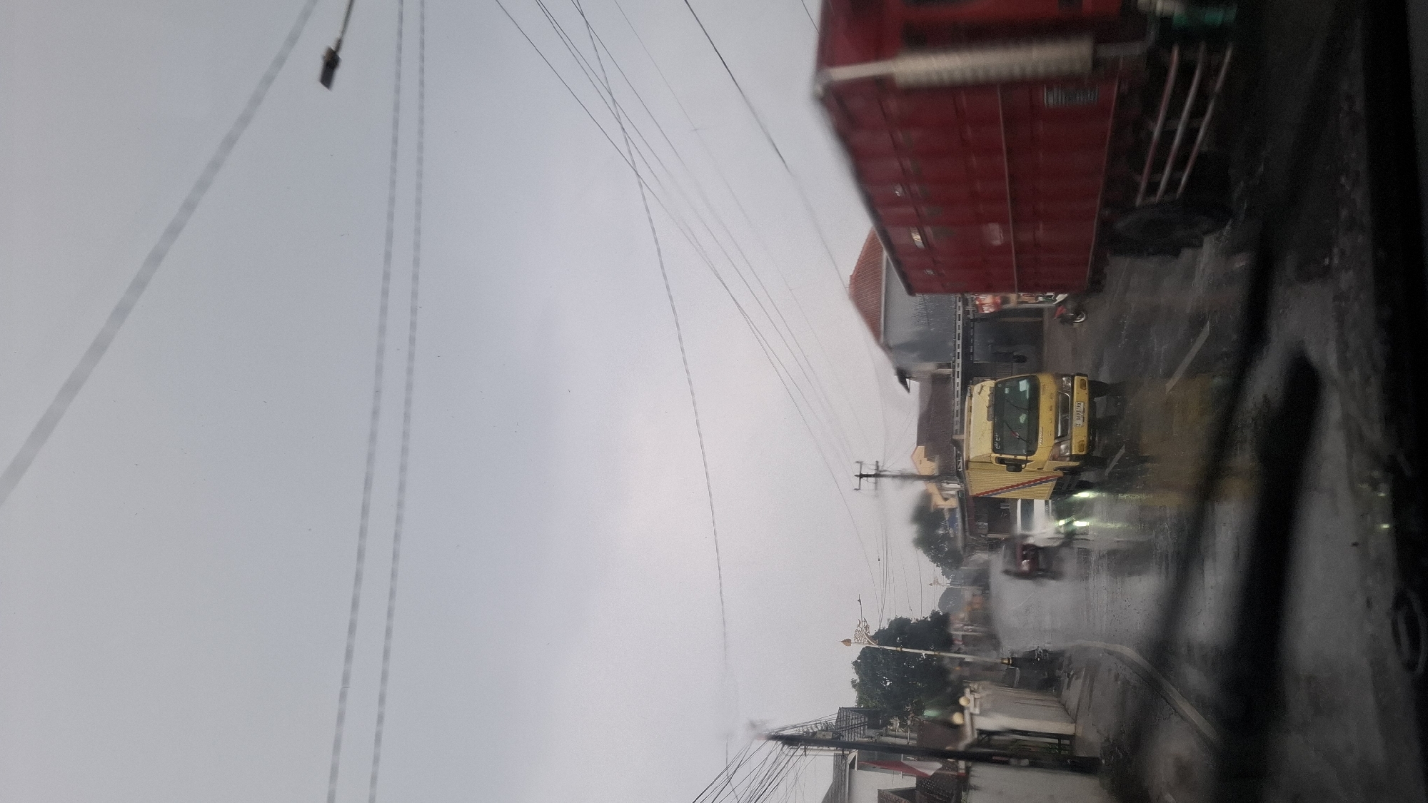 The image depicts a rainy street scene in Cilaku, Indonesia. In the foreground, the windshield of a vehicle is visible, with rain streaks and wiper marks. The road surface is wet and reflective, indicating recent rainfall. Several vehicles are on the road: a yellow truck with a white stripe on its side is in the middle lane, its headlights illuminated; a white car is to its left, also with headlights on; and a red truck with a blurry appearance is on the right side of the frame, partially obscuring the yellow truck. Further down the road, buildings line both sides, with visible rooftops and some architectural details. Utility poles and overhead wires are prominent, stretching across the frame. The sky is overcast and gray. A motorcycle is parked on the right side of the road. The overall impression is that of a busy road during inclement weather.