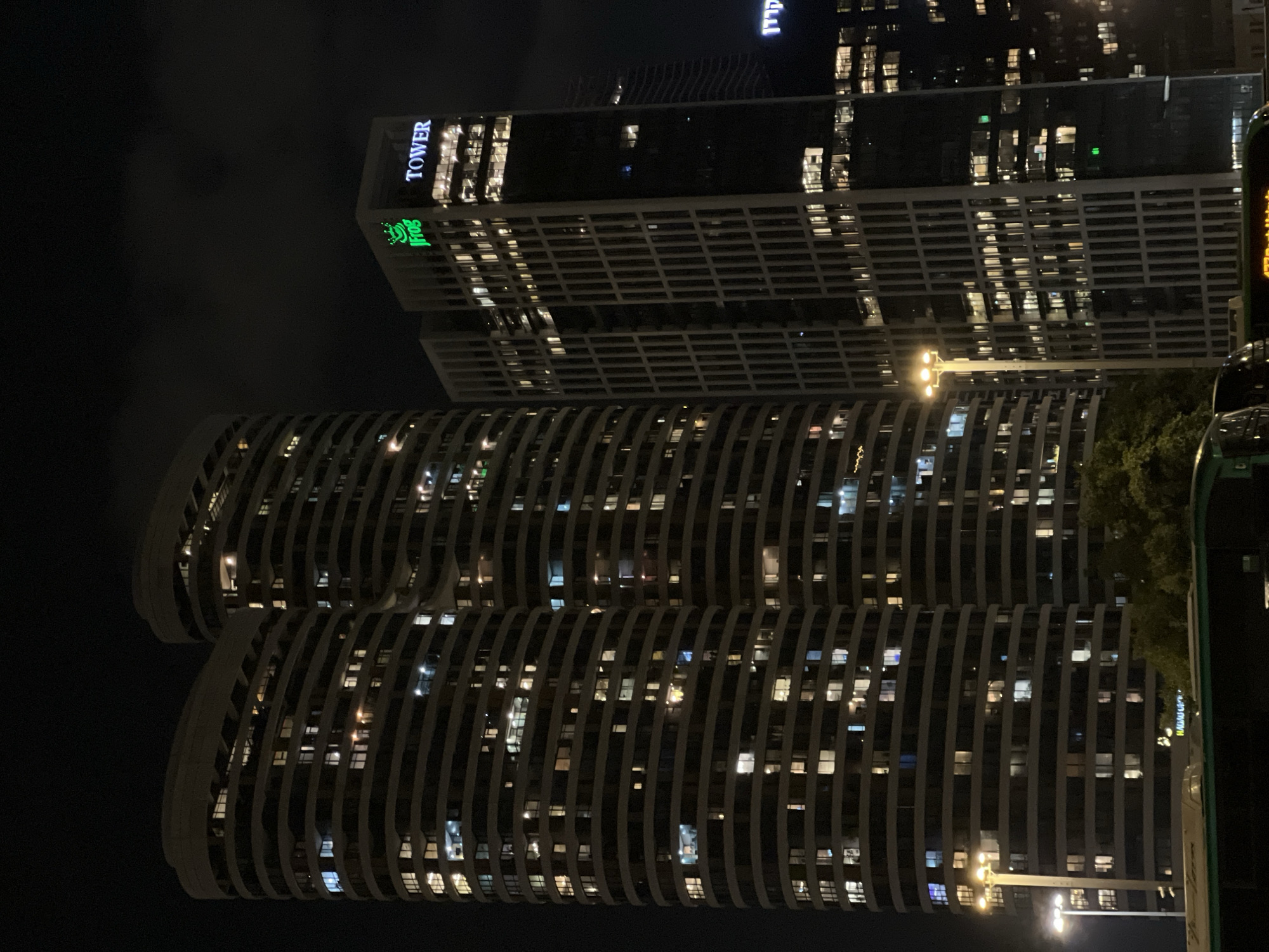 The image shows a nighttime view of modern skyscrapers in Tel Aviv, Israel. The prominent features are two large, curved, multi-story buildings with many illuminated windows, suggesting occupancy. To the right, a darker, rectangular tower stands taller, displaying a green neon sign with a frog logo and the text "Jfrog TOWER". Another building to the far right also has illuminated windows. In the foreground, the top of a green bus is visible, with a street lamp on the left side of the image illuminating the scene. The overall environment appears to be an urban setting with commercial or residential high-rise buildings.