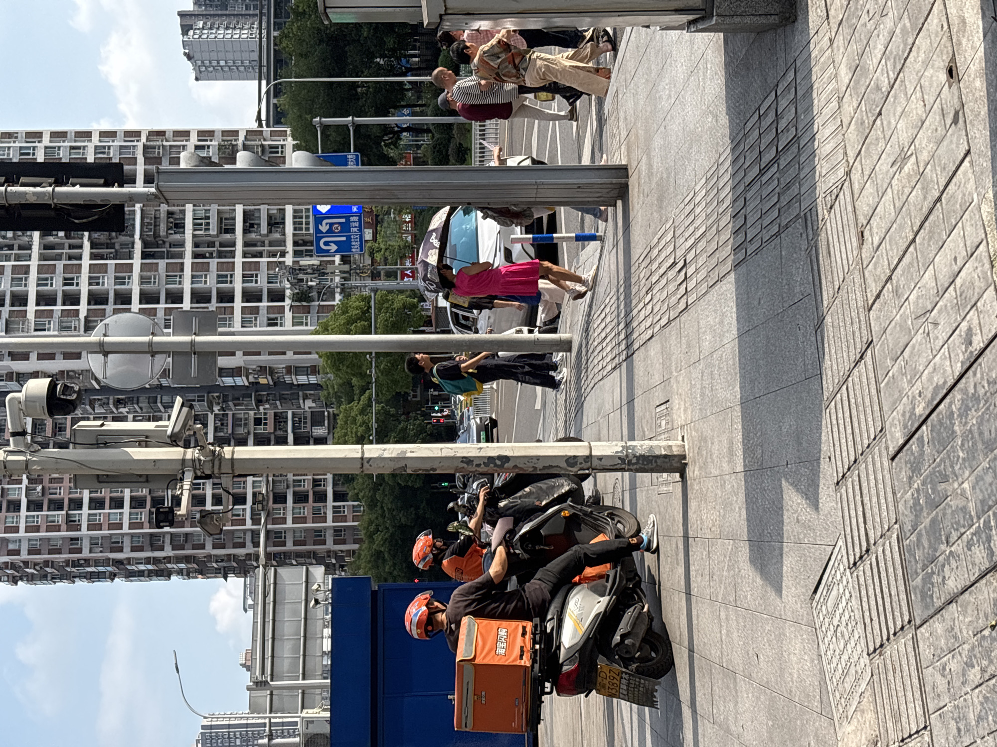 This is a street-level, eye-level, medium shot of a sunny day in Yuzhong District, China. In the foreground, a delivery driver wearing an orange helmet and a black shirt is seated on a white scooter. The scooter has an orange delivery box attached to the back with Chinese characters on it. To the right of the scooter, a sidewalk is visible with raised tactile paving for visually impaired pedestrians. Several people are walking on the sidewalk, including a woman in a pink dress holding an umbrella, and a group of individuals in casual clothing. In the background, tall apartment buildings with many windows dominate the skyline. Traffic lights and street signs are also visible, indicating a busy urban intersection. The overall impression is one of everyday urban life, with people going about their activities amidst the city's infrastructure.