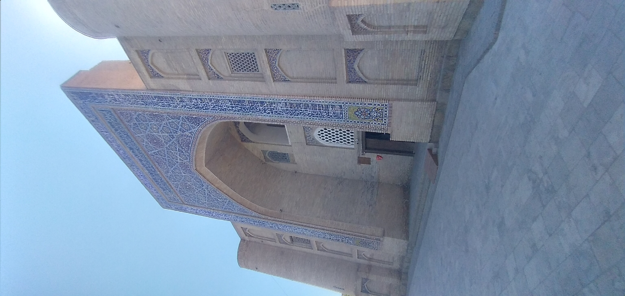 The image shows an upward-looking view of an intricately decorated building, likely a mosque or madrasa, with blue and white tilework and brickwork. The building features a large arched entrance adorned with elaborate geometric patterns and Arabic calligraphy in blue against a lighter background. Above the entrance, a band of blue tiles with white Arabic script is visible, bordered by smaller decorative tilework. The walls are constructed of light-colored bricks, with recessed niches and square windows featuring patterned screens. The sky, a clear light blue, occupies a significant portion of the image, suggesting it was taken on a sunny day. The ground in the foreground is paved with large, grey stone slabs laid in a grid pattern. The perspective is from a low angle, emphasizing the grandeur of the architecture. The overall impression is one of historical and religious significance, with a focus on ornate Islamic art and design. No people are visible in the frame. The time of day appears to be daytime, given the bright sky.