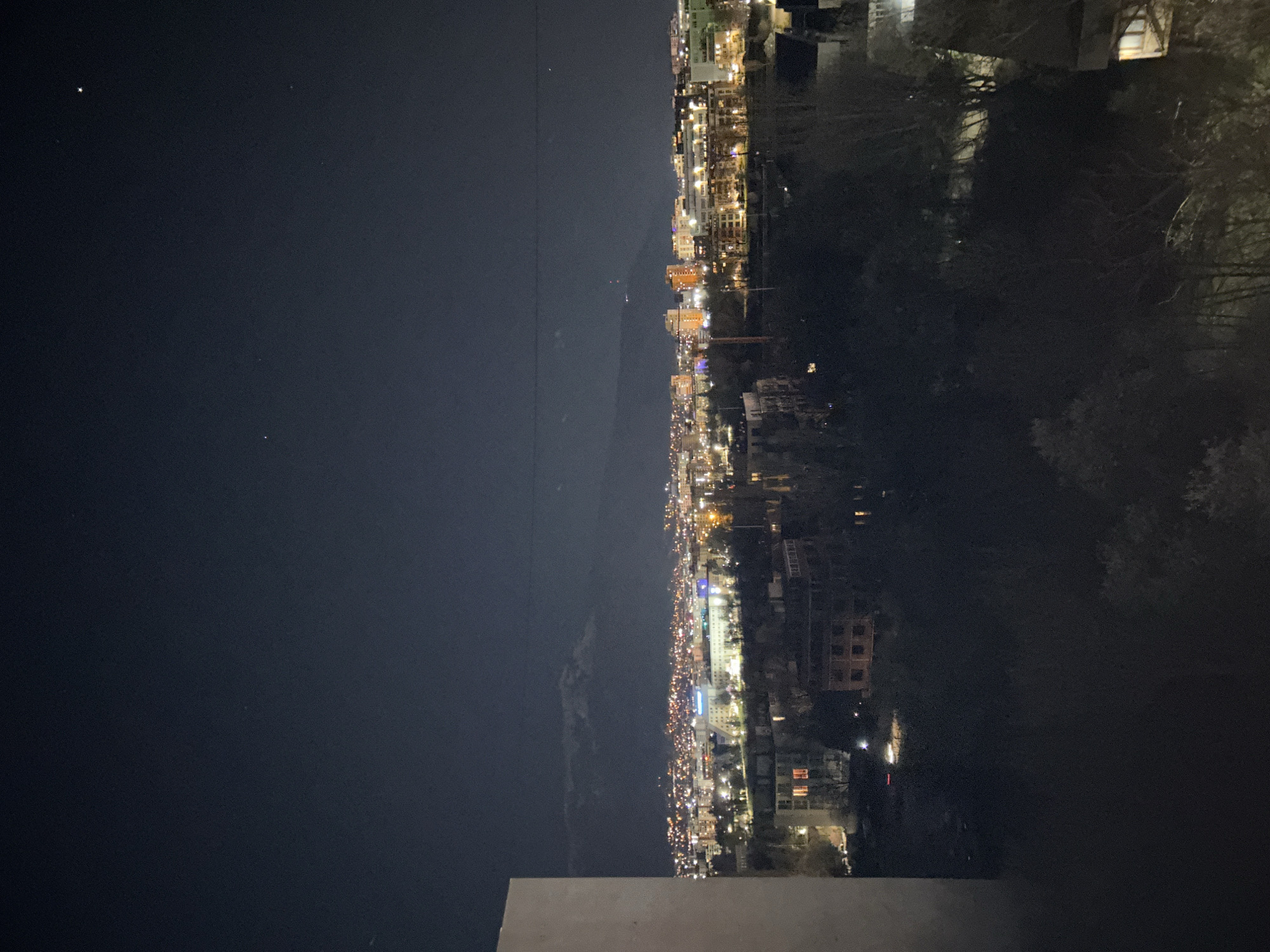 A nighttime shot shows a city skyline. On the right, trees are visible, partially obscuring buildings with lit windows. The city lights illuminate the buildings. On the left, the night sky is dark with a few visible stars. There appear to be mountains in the distance. The city is the city of Sofia, Bulgaria.