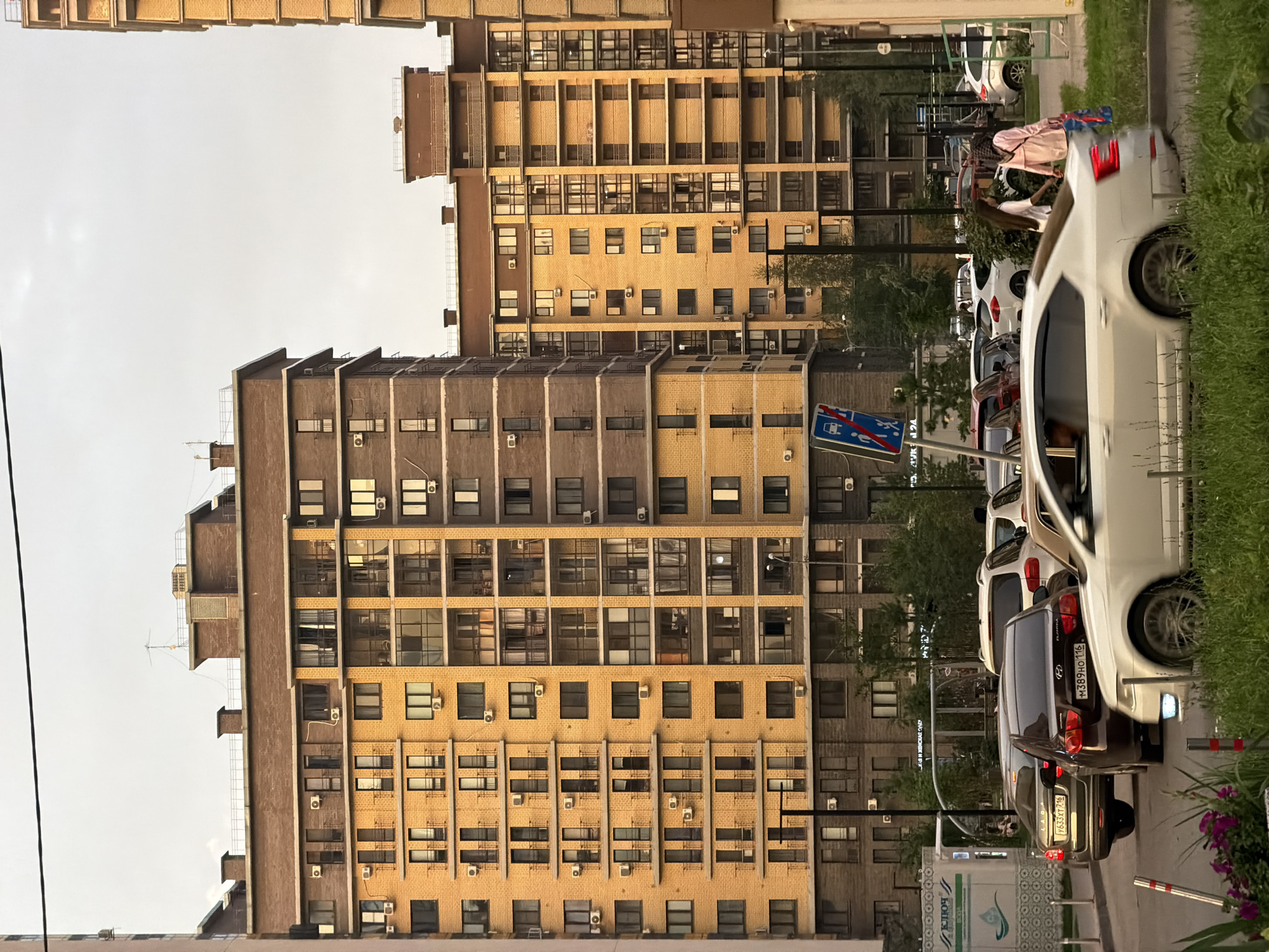 The image depicts an urban scene in Kazan, Russia, focusing on a street in front of modern apartment buildings. Two tall apartment buildings, one with a yellow brick facade and the other with a darker brick facade, dominate the background. The yellow building has numerous windows and balconies, many of which are illuminated, suggesting it is late afternoon or evening. Several cars are parked and driving on the street. In the foreground, a white sedan is moving, and to its right, two women are walking. One woman wears a pink jacket and the other a white shirt and dark pants. A street sign indicating a pedestrian crossing and a prohibition is visible on the left side of the street. The sky is overcast, creating a soft light. The street is lined with trees and grass, and there are visible air conditioning units on the buildings.