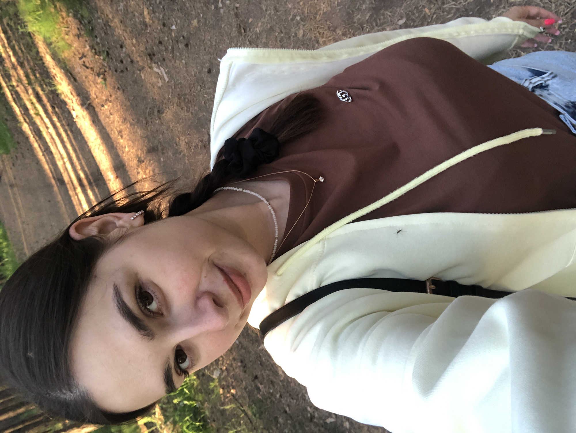A young woman is taking a selfie outdoors. She has dark hair pulled back in a braid with a black scrunchie and is wearing a brown t-shirt, a cream-colored hoodie with yellow drawstrings, and light-wash jeans. She is wearing multiple necklaces and a jewel earring. The background appears to be a wooded area with light filtering through the trees onto a dirt path. Based on the presence of Russian text in the image metadata, it is likely taken in the city of Chaikovsky, Russia.