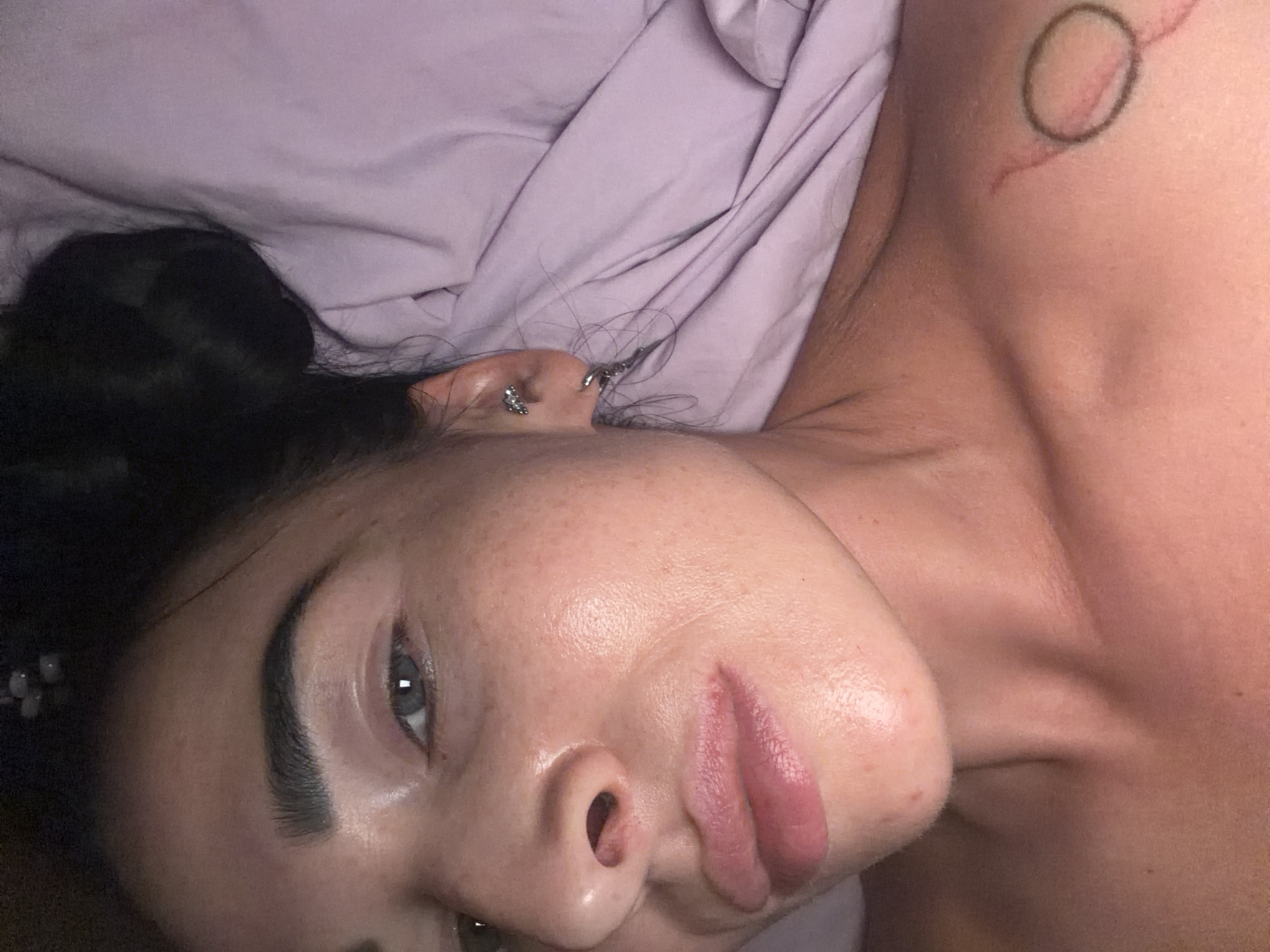 A close-up, portrait-style shot shows a person lying down on their side, facing the camera. The subject is a young woman with dark hair, which appears to be pulled back in a bun, and fair skin with visible freckles across her nose and cheeks. She has distinct, well-groomed dark eyebrows and blue or grey eyes. Her lips are full and have a natural, soft pink hue.
She is wearing multiple ear piercings: a silver-toned earring resembling a butterfly or floral design in her helix, and another silver-toned ring or stud in her lobe. A subtle, circular tattoo is visible on her right shoulder or upper chest area, accompanied by a faint reddish mark.
The background consists of light purple or lavender bedding, possibly a sheet or duvet, which is slightly rumpled. The lighting is soft, suggesting an indoor setting, possibly morning or evening. The overall mood is relaxed and calm. No specific objects or text are visible in the background that would indicate a particular location or activity beyond resting. There are no visible location cues within the image itself to suggest Saint Petersburg, Russia.