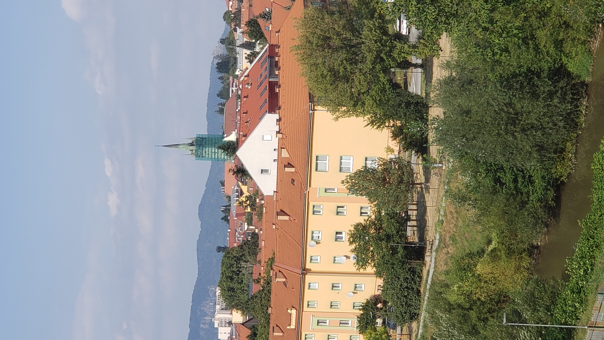 The image captures a daytime, elevated view of a residential area in Prešov, Slovakia. In the foreground, dense green foliage lines a calm body of water. Beyond the vegetation, a row of apartment buildings with orange tiled roofs dominate the midground. These buildings are characterized by their light yellow facades and numerous windows. A tall, green-clad spire of a church or similar structure is visible in the background, rising above other buildings and a range of blue-hued hills. The sky is a clear blue with scattered white clouds. The scene is devoid of people.