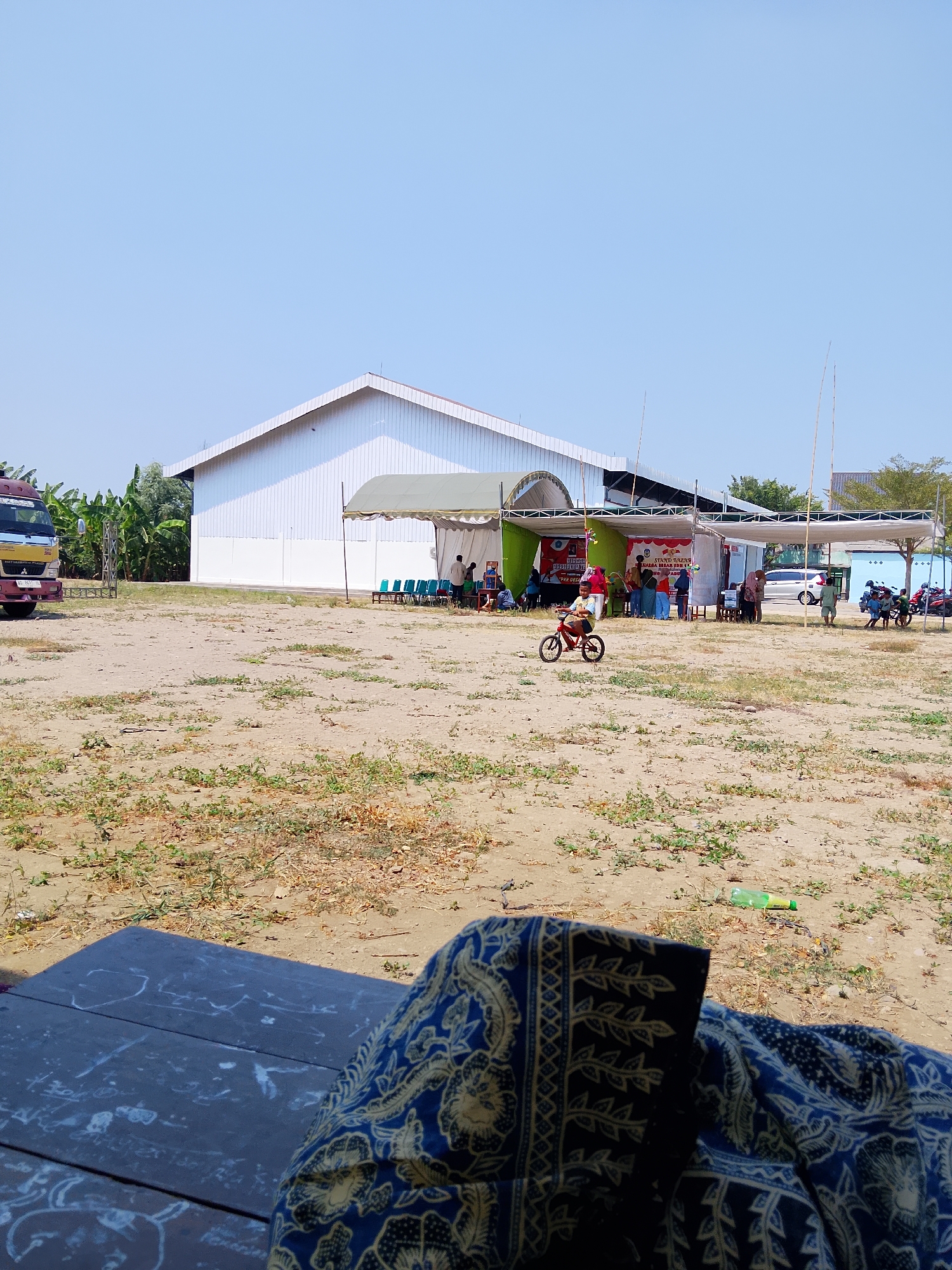In Pelem, Indonesia, an outdoor event is taking place under a clear blue sky. A large, white, corrugated metal building with a pitched roof serves as a backdrop. In front of the building, several white tents are erected. Under the tents, there are rows of turquoise chairs and various stalls with signage, suggesting a bazaar or exhibition. People are gathered around the stalls, interacting and observing.

In the foreground, a young child is riding a red bicycle. The ground is mostly bare earth with sparse patches of dry grass and scattered debris, including a green plastic bottle. A dark-colored table with white markings is visible in the lower portion of the image, draped with a blue and yellow patterned fabric. To the left, a truck with red and yellow detailing is parked. Banana trees and other greenery are visible behind the truck and to the left of the building. The overall scene depicts a lively community event in an open, somewhat dusty environment.