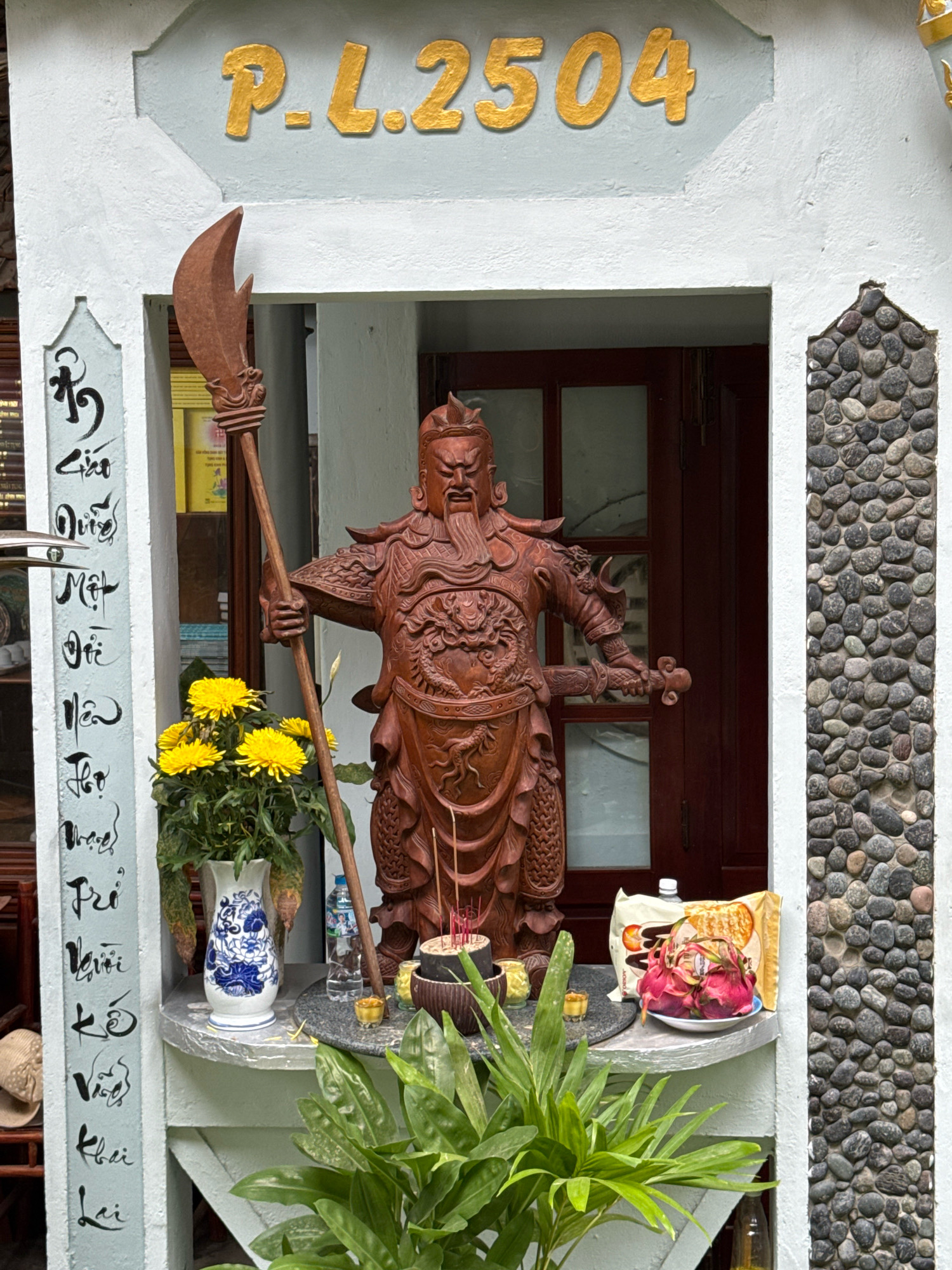 A wooden statue of a warrior with a beard and armor stands in an alcove. The warrior holds a spear in its right hand and a sword in its left. In front of the statue, there is a vase of yellow flowers, incense sticks in a pot, and offerings of dragon fruit and packaged snacks. The alcove is decorated with stone pebbles on the right side and calligraphy on a plaque to the left. Above the alcove, gold lettering reads "P.L. 2504". The scene appears to be a shrine or altar. The architectural style and the golden lettering with what appears to be Vietnamese characters suggests the location is in Vietnam, likely a residential area or a temple in Nha Trang, given the context.