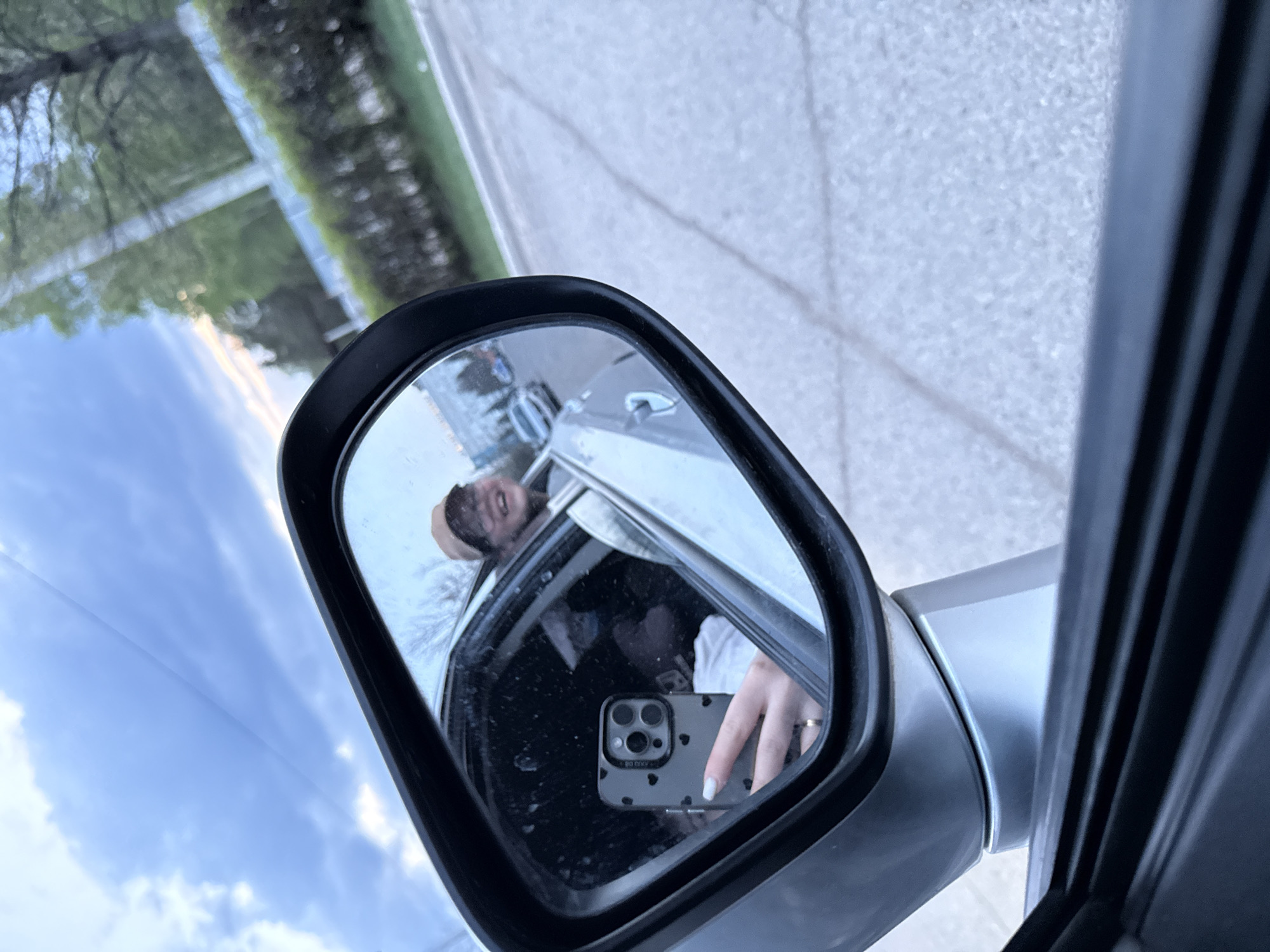 The image shows a selfie taken in a car's side mirror. The reflection shows a person holding a phone with a case decorated with black hearts. The person appears to be smiling. Outside the car, a road and trees are visible. There is no evidence of the location being in Chaikovsky, Russia.