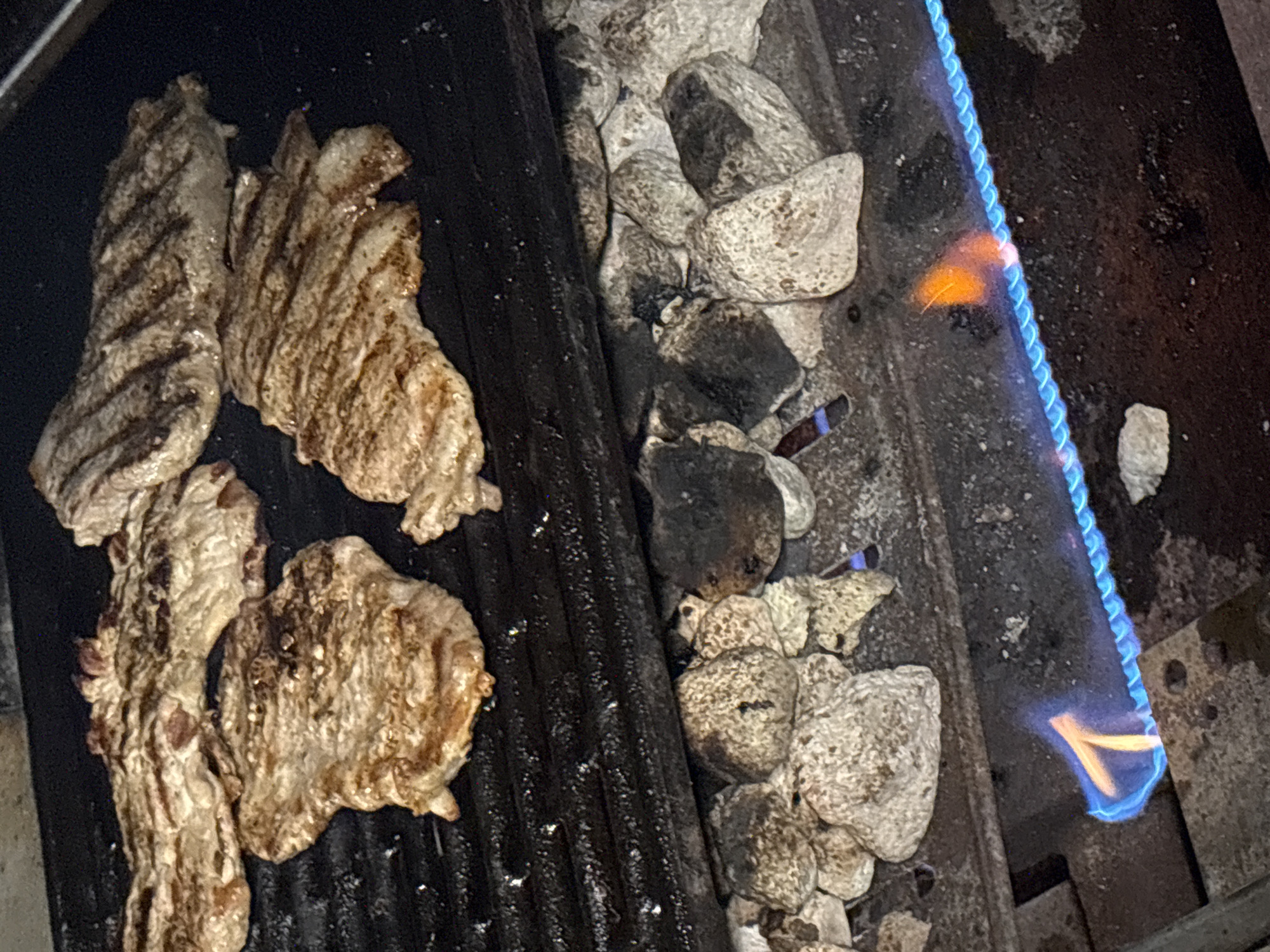 The image shows a close-up of a grill. Four pieces of meat are cooking on the grill grates. To the right of the meat are ceramic briquettes. A blue and orange flame is visible on the right side of the image, likely from a gas burner. The grill appears to be outdoors, but the surrounding environment is not visible. There is no indication of the location being in Lehchevo, Bulgaria, or any information about people or specific actions.