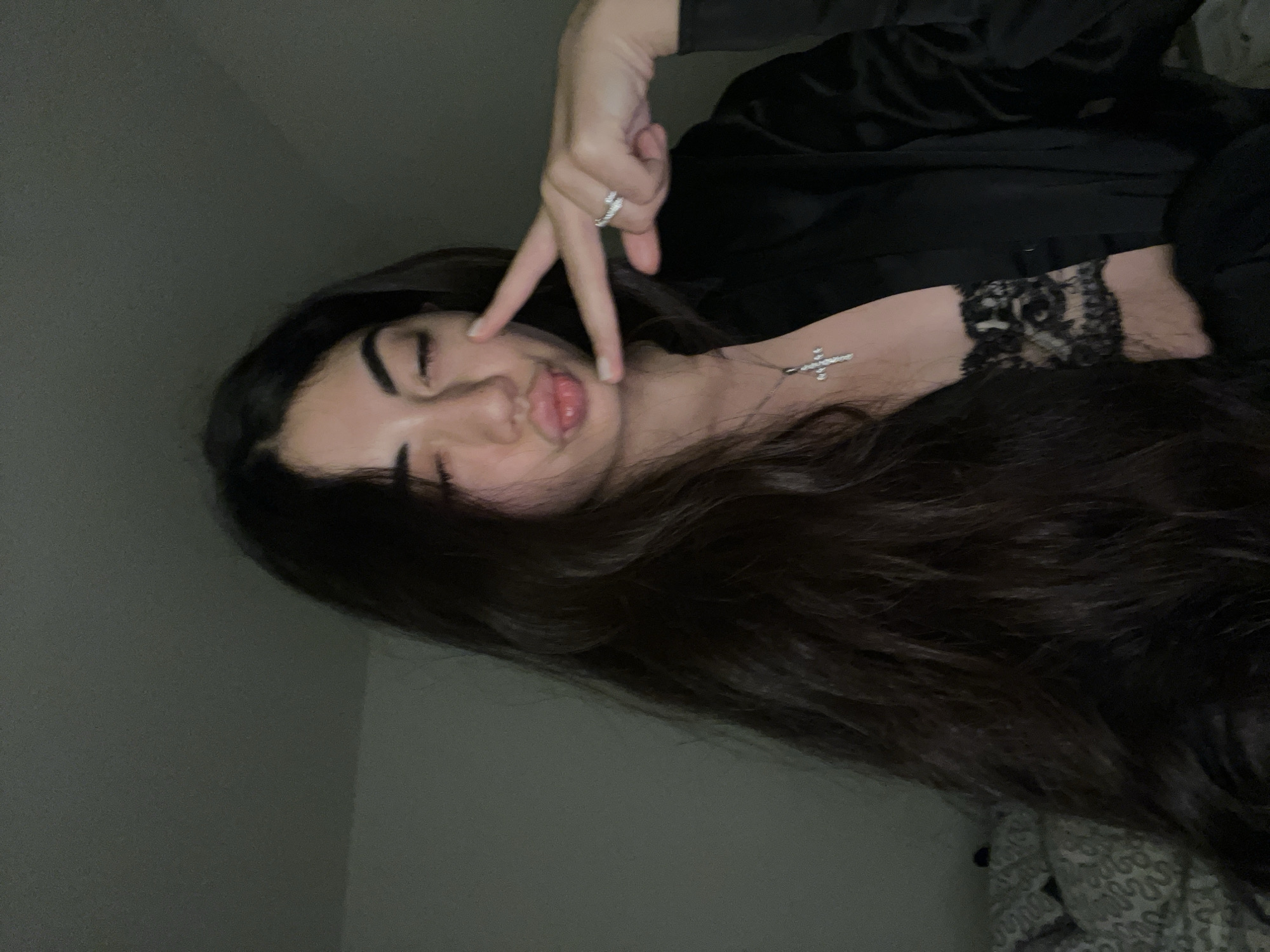 The image shows a young woman with long, dark hair posing with her eyes closed and lips puckered. She is wearing a black satin robe over a black lace bra and a silver cross necklace. Her right hand is held up, displaying a peace sign and a ring on her finger. The background is a dark, nondescript room, suggesting an indoor setting. The image is a close-up selfie. There is no visible information in the image that would suggest it was taken in the city of Chotěšov, Czechia.