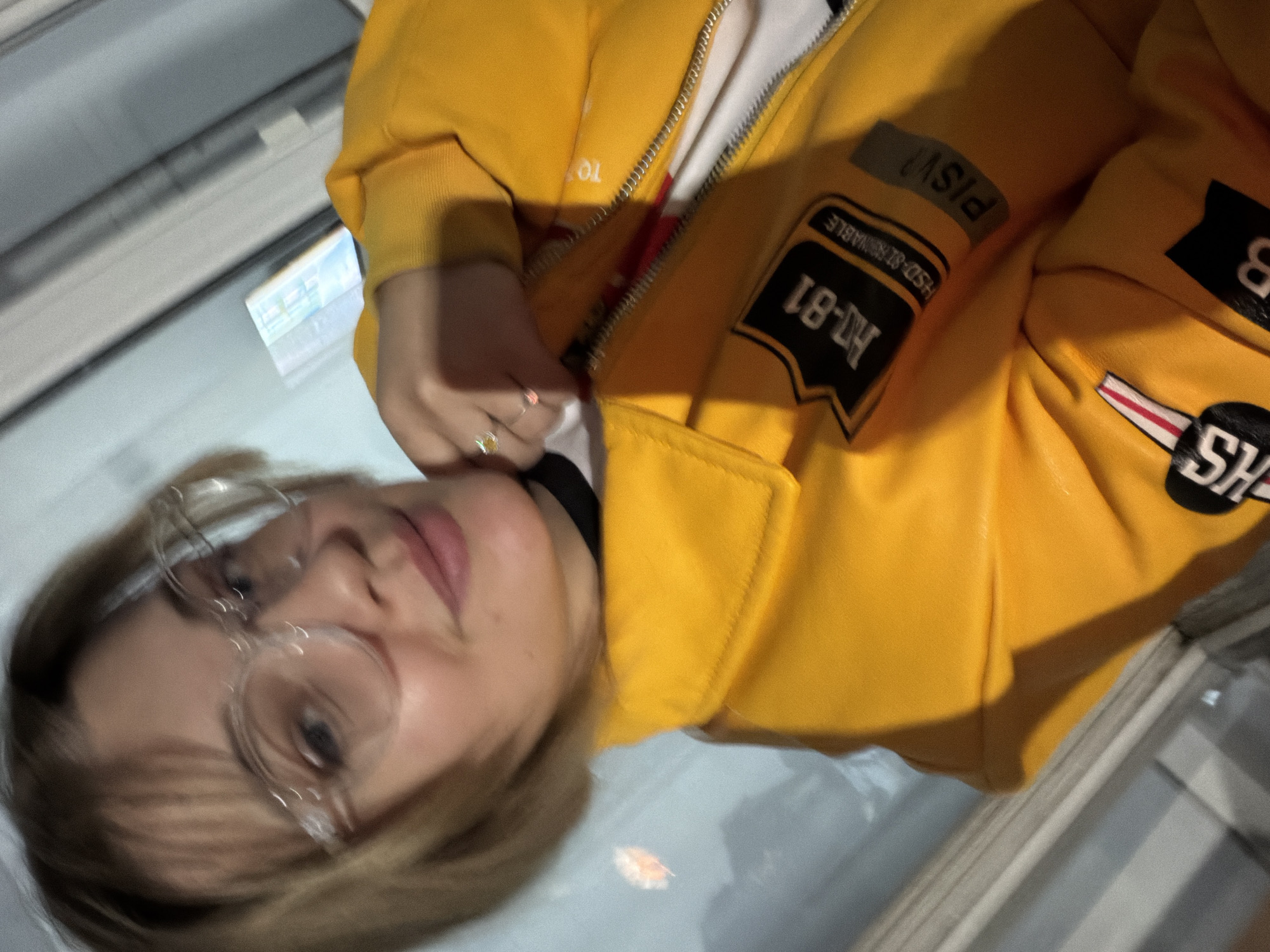 A young woman with blonde hair and clear glasses is seen close-up, wearing a yellow jacket with patches. The jacket has a zipper being pulled up by her left hand, which is adorned with a ring. The patches on the jacket have lettering and numbers, including "H-81" and "SH". The background suggests an indoor environment, possibly near a window with a blurry reflection. The photo was taken in Kazan, Russia.