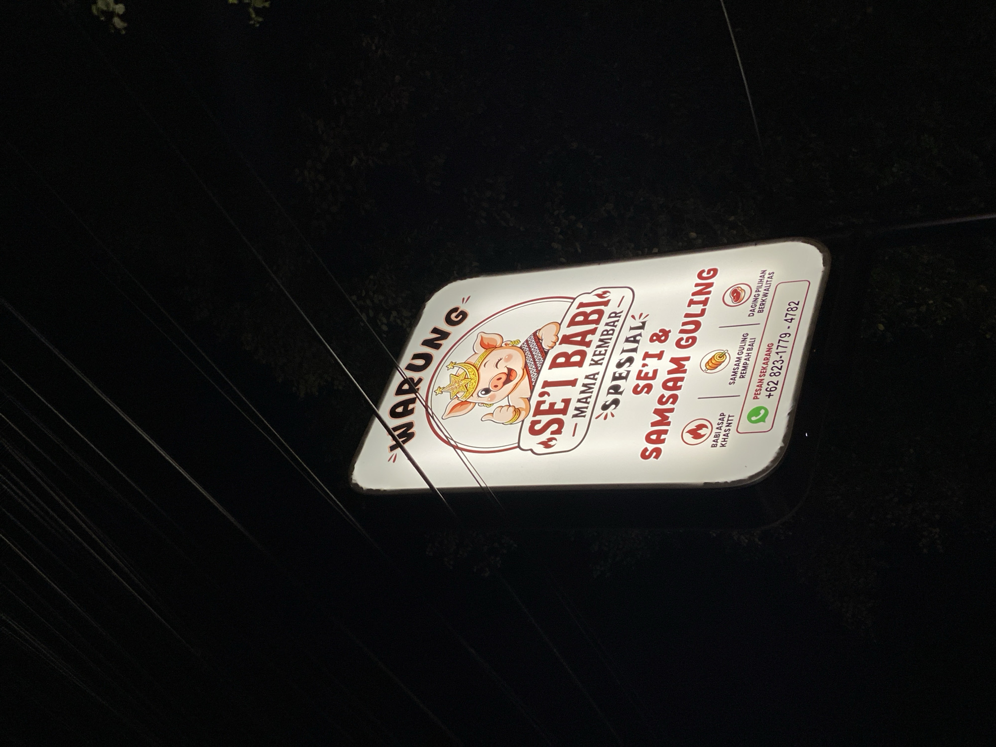 The image shows a lit-up sign for "WARUNG SE'I BABI - MAMA KEMBAR" in Kuta Selatan, Indonesia. The sign features a cartoon pig wearing a crown and giving a thumbs up. Below the pig, the text advertises "SPESIAL SE'I & SAM SAM GULING". There are also icons representing "BABI ASAP KHAS NTT", "SAM SAM GULING REMPAH BALI", and "DAGING PILIHAN BERKUALITAS". A WhatsApp contact number, "+62 823-1779-4782", is displayed at the bottom. The sign is illuminated against a dark, outdoor background, with power lines visible in the upper part of the image. The overall lighting suggests it is nighttime.
