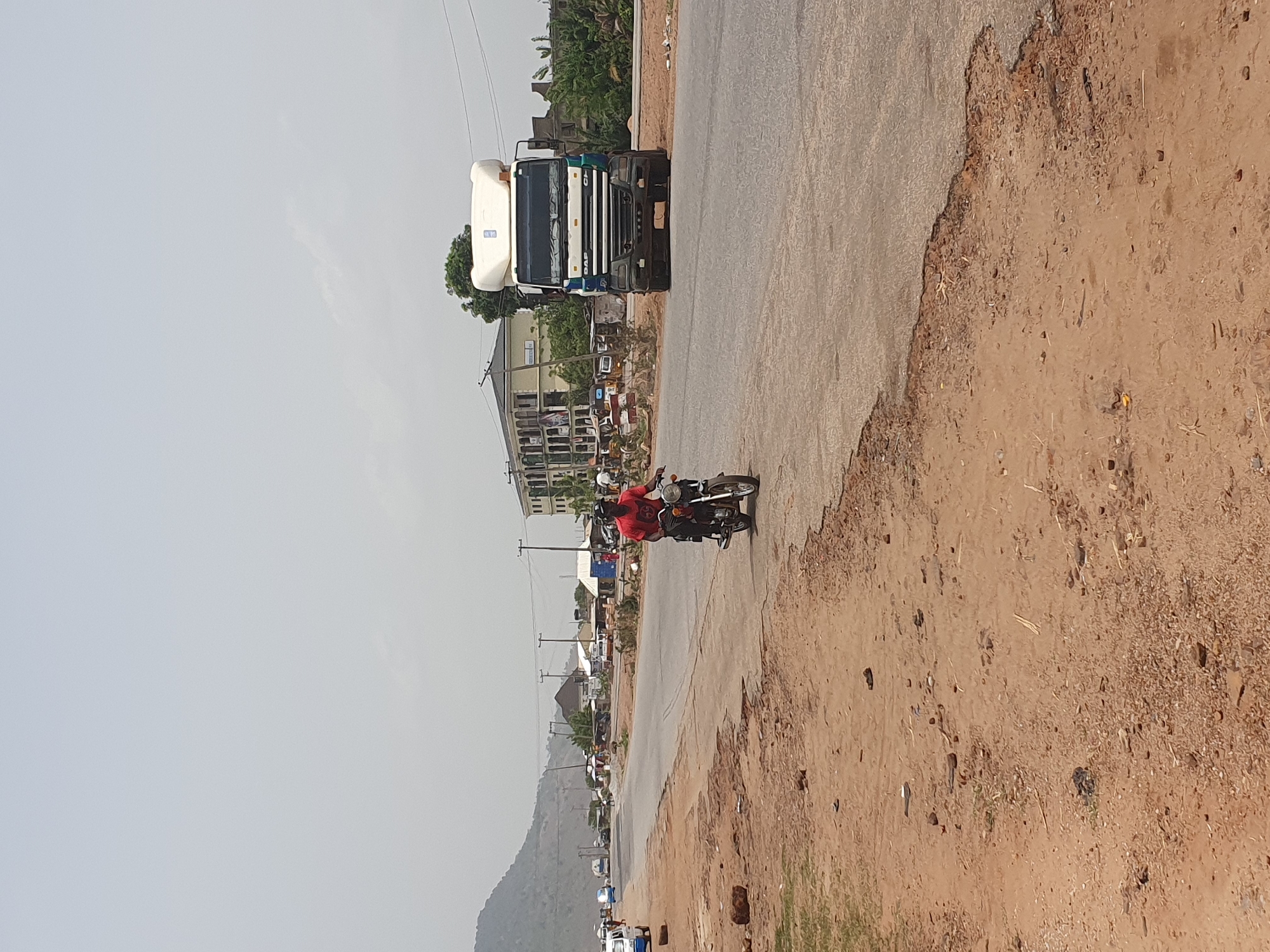 The image, rotated 90 degrees clockwise, depicts a daytime scene on a semi-urban or rural road in Lokoja, Nigeria. A paved road stretches across the frame, bordered by a rough, sandy, and unpaved shoulder on its right. In the midground, a person wearing a red t-shirt is riding a dark-colored motorcycle on the paved section of the road.
Further down the road, on the left side, a large white and blue semi-trailer truck is visible, either parked or moving slowly. The brand name "DAF" is clearly visible on the front grille of the truck. Along the roadside, various buildings, which appear to be a mix of residential and small commercial structures, line the street. Several other vehicles, including cars and motorcycles, are discernible in the distance, suggesting moderate traffic.
In the far background, a range of hills or mountains rises under a light, hazy, or overcast sky. The road surface shows signs of wear, particularly where the asphalt meets the dirt shoulder, indicating some disrepair. The scene captures a typical day on a thoroughfare in the region.