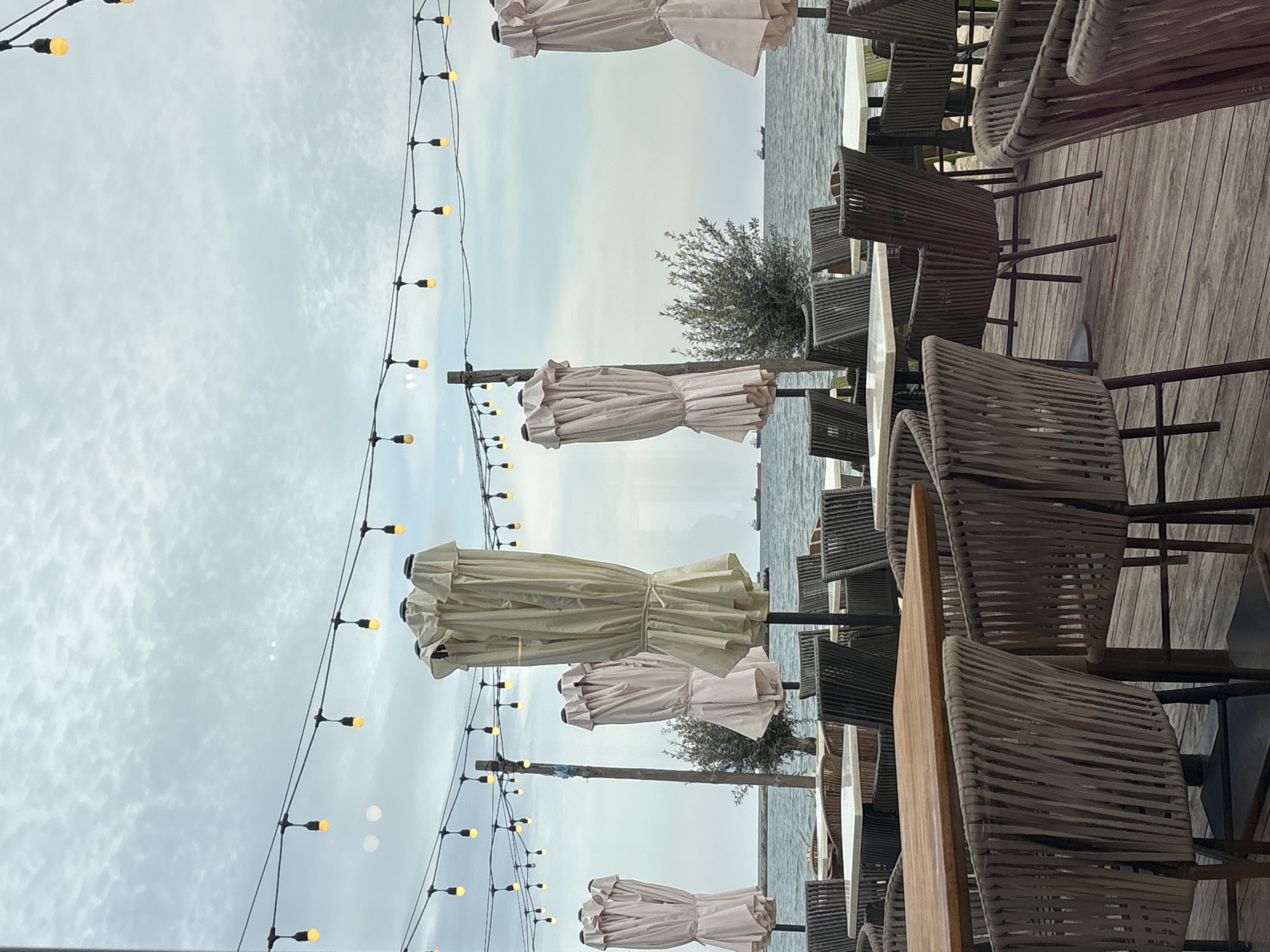 The image depicts an outdoor dining area in Varna, Bulgaria, situated near the sea. Several closed umbrellas are positioned along a line, with strings of lights hanging between them. Tables with chairs surround the umbrellas, suggesting a restaurant or cafe. In the background, the sea extends to the horizon under a cloudy sky. The scene is composed with a vertical orientation, and the image shows a mix of natural and artificial lighting. No people are visible in the image.