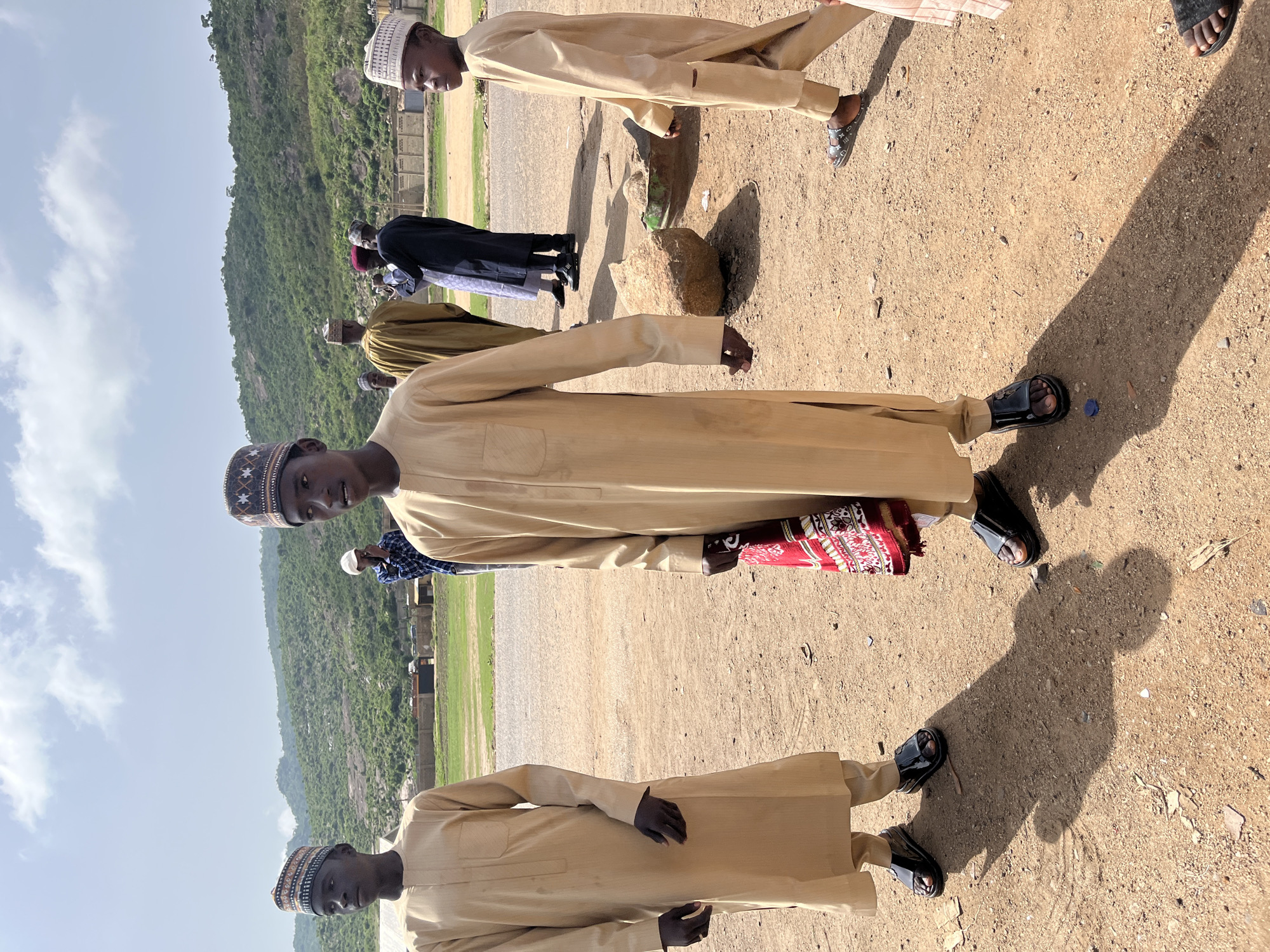 In Naraguta Hausawa, Nigeria, a group of men are gathered outside. The men are wearing traditional Hausa attire, including embroidered caps. The landscape includes a mountain and an open field. The ground is sandy. The men are standing in a row, some holding prayer mats. Some men are wearing sandals. Other people are in the background.