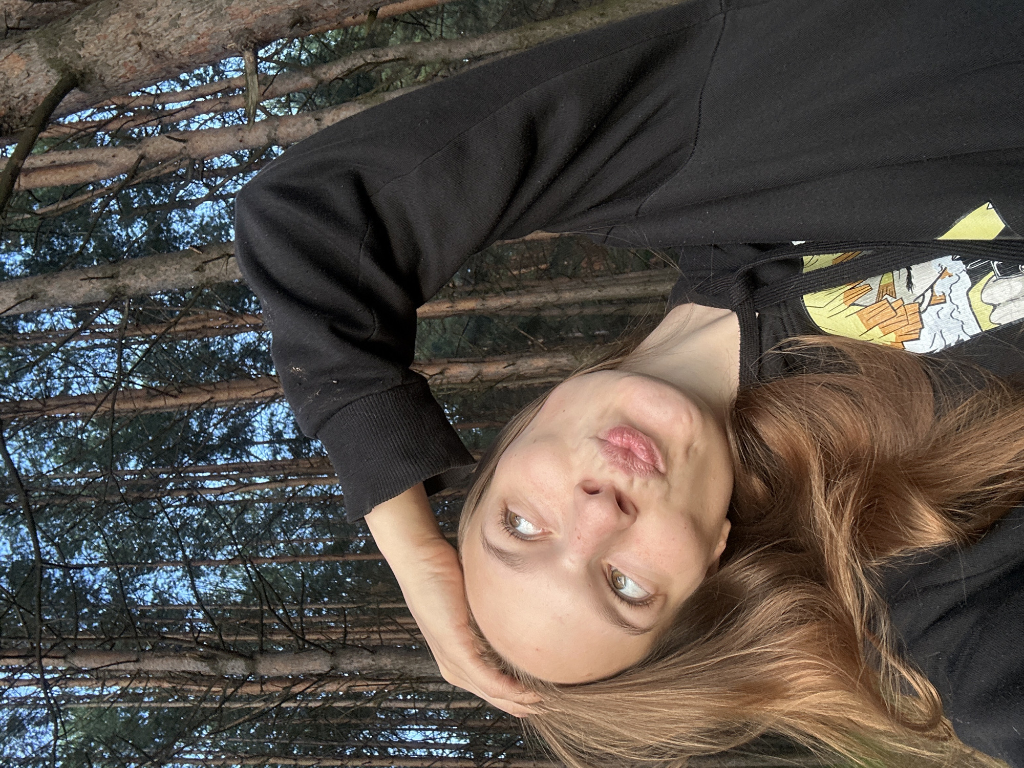 A young woman with long, light brown hair is taking a selfie outdoors in a forest. She is wearing a black hoodie with a graphic print on the left side. Her head is tilted back, and her left hand is resting on her forehead. Her eyes are looking upwards, and her lips are pursed in a pout. The background consists of numerous tall pine trees with a clear blue sky visible through the branches. The image appears to have been taken during the daytime, possibly in the late afternoon or early evening, given the lighting. The location is a forest in the city of Chaikovsky, Russia.