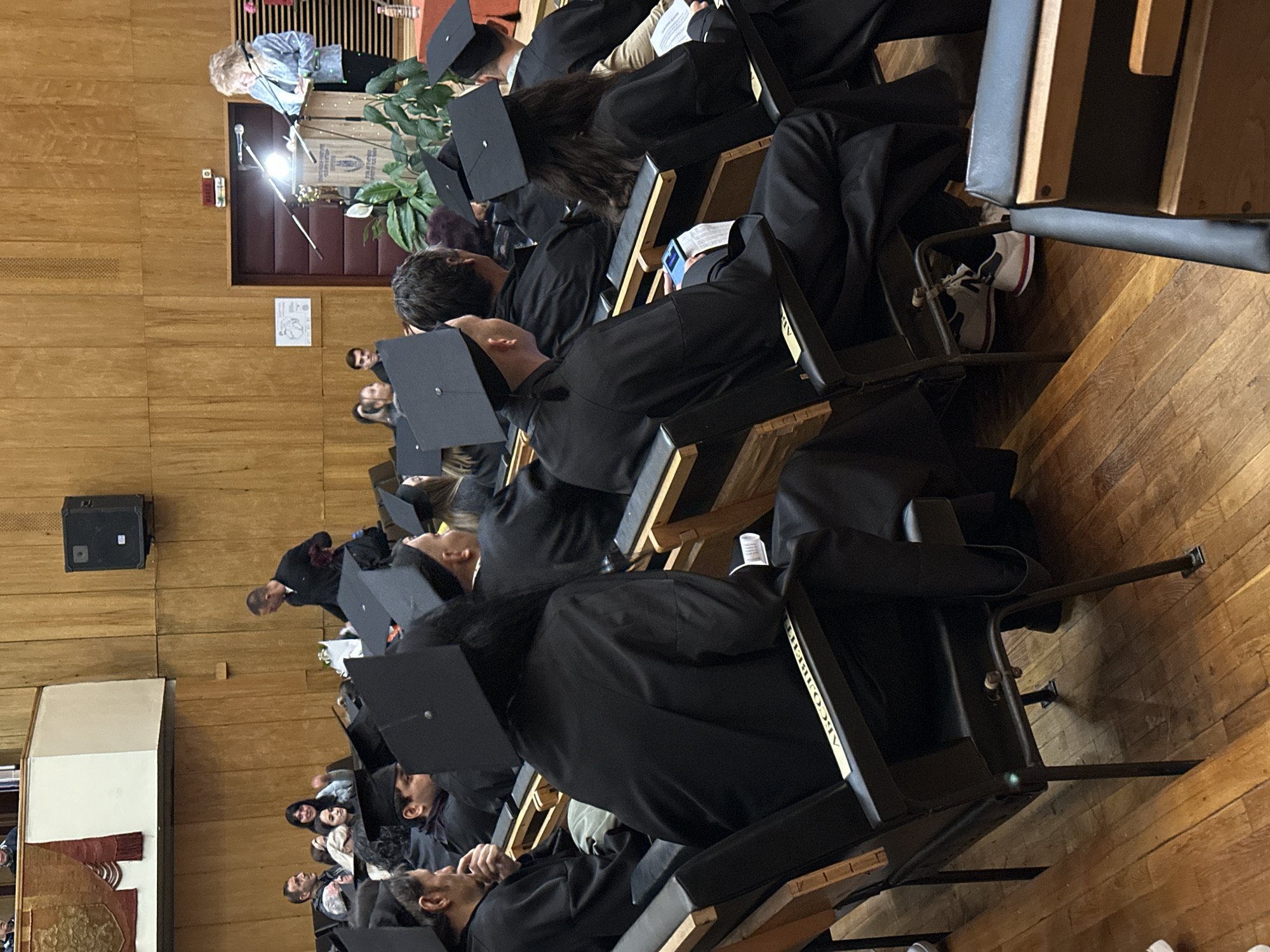 The image shows an indoor graduation ceremony in a room with wood-paneled walls and a wooden floor. Rows of graduates wearing black gowns and mortarboards are seated in black chairs. A woman is speaking at a podium, positioned in front of a door. A speaker is mounted on the wall. The location is likely Veliko Tarnovo, Bulgaria, although this cannot be definitively determined from the image alone. The students are listening, with some appearing to be distracted or looking at their phones.
