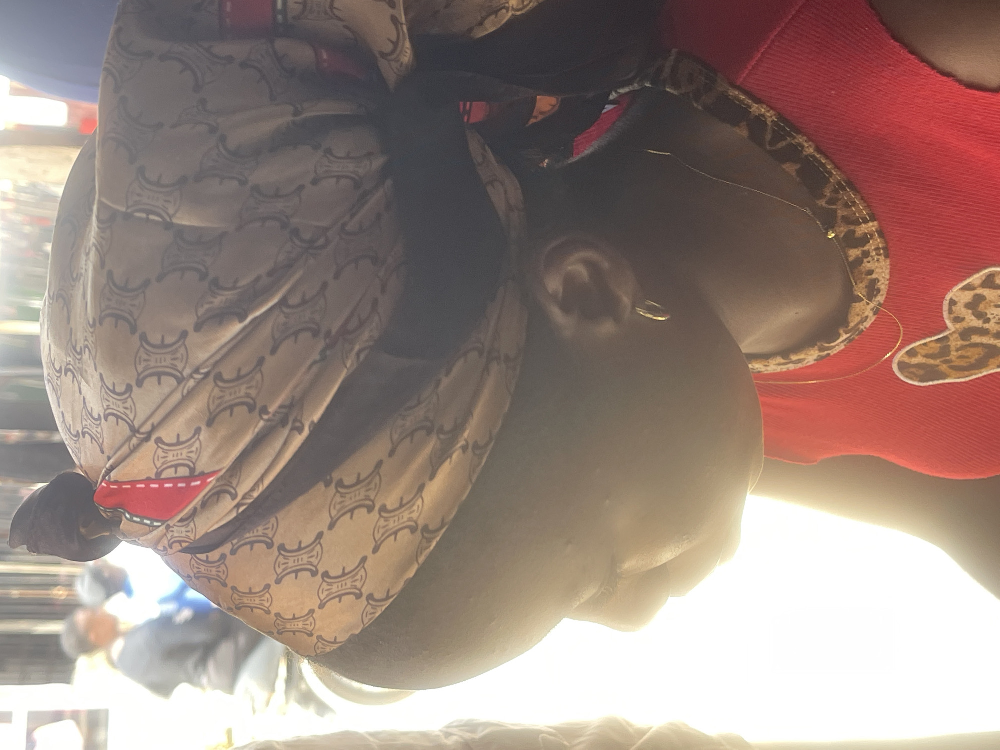 The image shows a close-up profile view of a young Black female wearing a headscarf with a repeating "C" logo pattern in brown and black. A red fabric panel is visible on the headscarf. She is wearing a red sleeveless top with a leopard print trim around the neckline and a delicate gold necklace. A hoop earring is visible in her left ear. The background is blurred, showing people and what appears to be market stalls or shops, suggestive of an outdoor setting in Abuja, Nigeria. The lighting is bright and somewhat overexposed, particularly on the left side of the image, creating a strong silhouette effect.