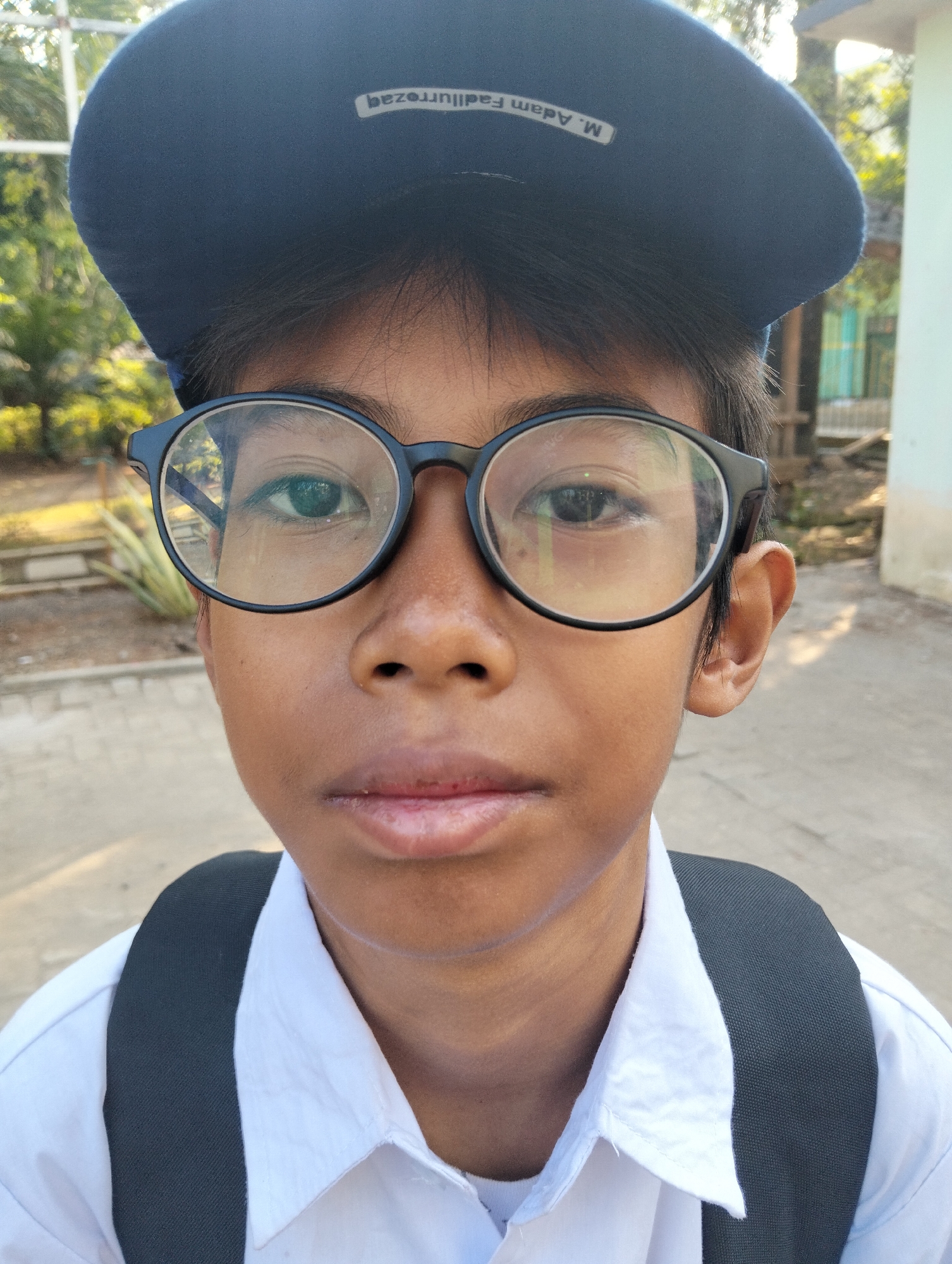 A close-up, head-and-shoulders shot features a young person wearing a dark blue cap and black-rimmed glasses, facing the camera. The person has dark hair, visible beneath the cap, and brown skin. They are wearing a white collared shirt, with the straps of a dark backpack visible over the shoulders. The cap has a white embroidered text that reads "M. Adam Fadillurrezq". The background is out of focus, showing green foliage, a pale wall, and a paved ground, suggesting an outdoor urban or semi-urban setting. The lighting is bright, indicating daylight. The overall impression is that of a candid portrait taken outdoors, possibly near a school or residential area, in the city of Paciran, Indonesia.