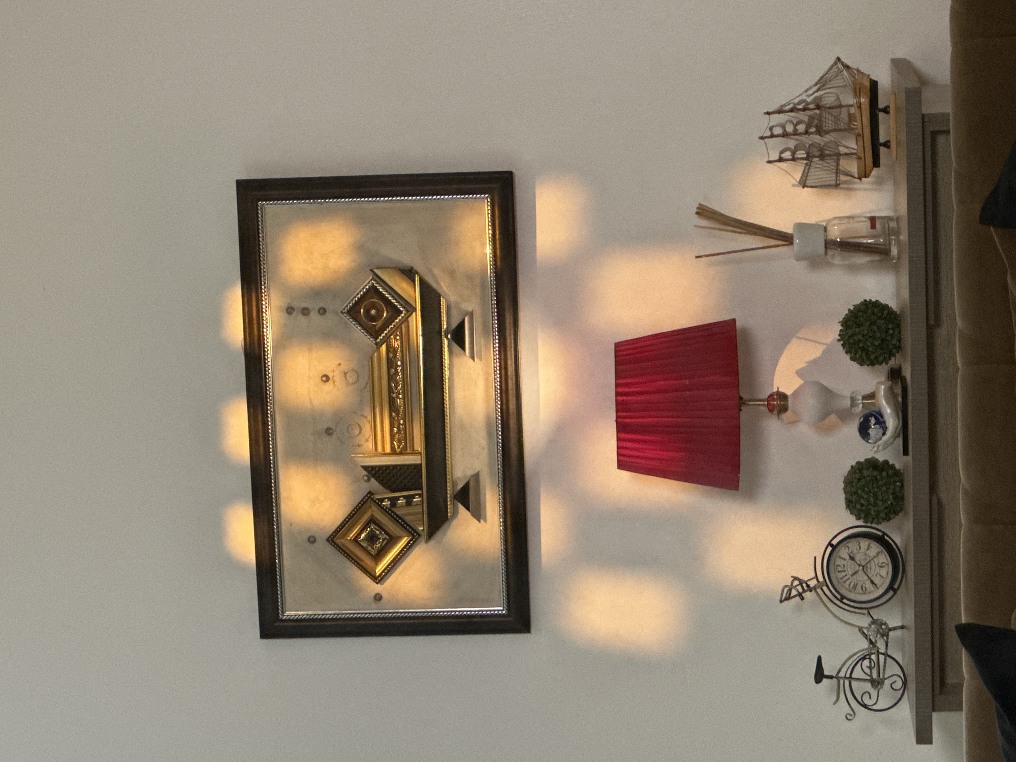 The image depicts an indoor scene, likely a living room, with a wall adorned by a rectangular framed artwork. The artwork features a geometric design with metallic accents in gold and silver, reflecting the warm light of what appears to be sunset or artificial lighting. Below the artwork, on a narrow shelf or mantle, several decorative objects are arranged.
From left to right, these objects include a miniature metal bicycle, a clock designed to resemble a bicycle wheel, a small green spherical plant, a white and red table lamp with a pleated shade, a diffuser with reeds, and a model of a wooden sailing ship. The shelf is positioned above a piece of brown furniture, possibly a sofa.
The lighting creates a dramatic effect, with distinct rectangular shadows cast on the wall above and to the left of the shelf, originating from an unseen light source or window. The overall ambiance is one of a decorated interior space with a focus on decorative items and the play of light and shadow.
While the image is detailed, it does not contain any visual information that allows for the specific identification of its location within Pakuro, Nigeria. The scene is generic to many interior decorations and lacks any identifying geographical markers or cultural specificities of the region.