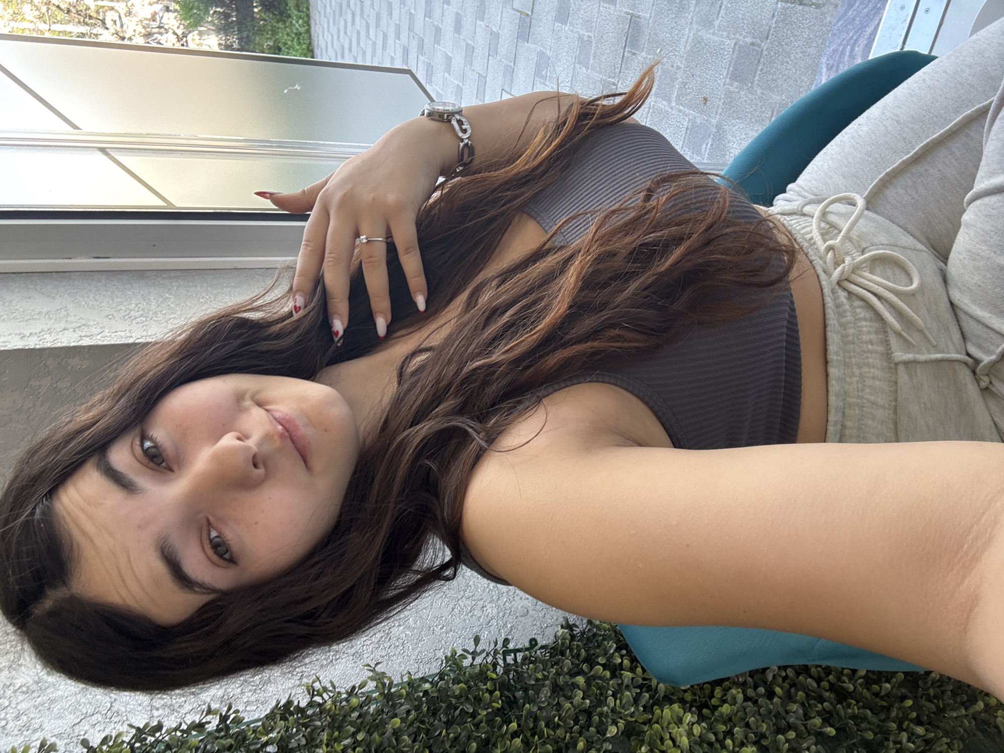 A young woman with brown hair and fair skin is positioned in the foreground, likely taking a selfie. She is wearing a grey ribbed tank top and light grey sweatpants. A silver watch adorns her left wrist, and a silver ring is on her finger. Her fingernails appear to have a design, with one nail showing a small red heart.
She is seated on a teal cushion, which is placed on artificial greenery. The background features a sliding glass door and a light grey brick wall. The location details in Plovdiv, Bulgaria, are not discernable in the image.