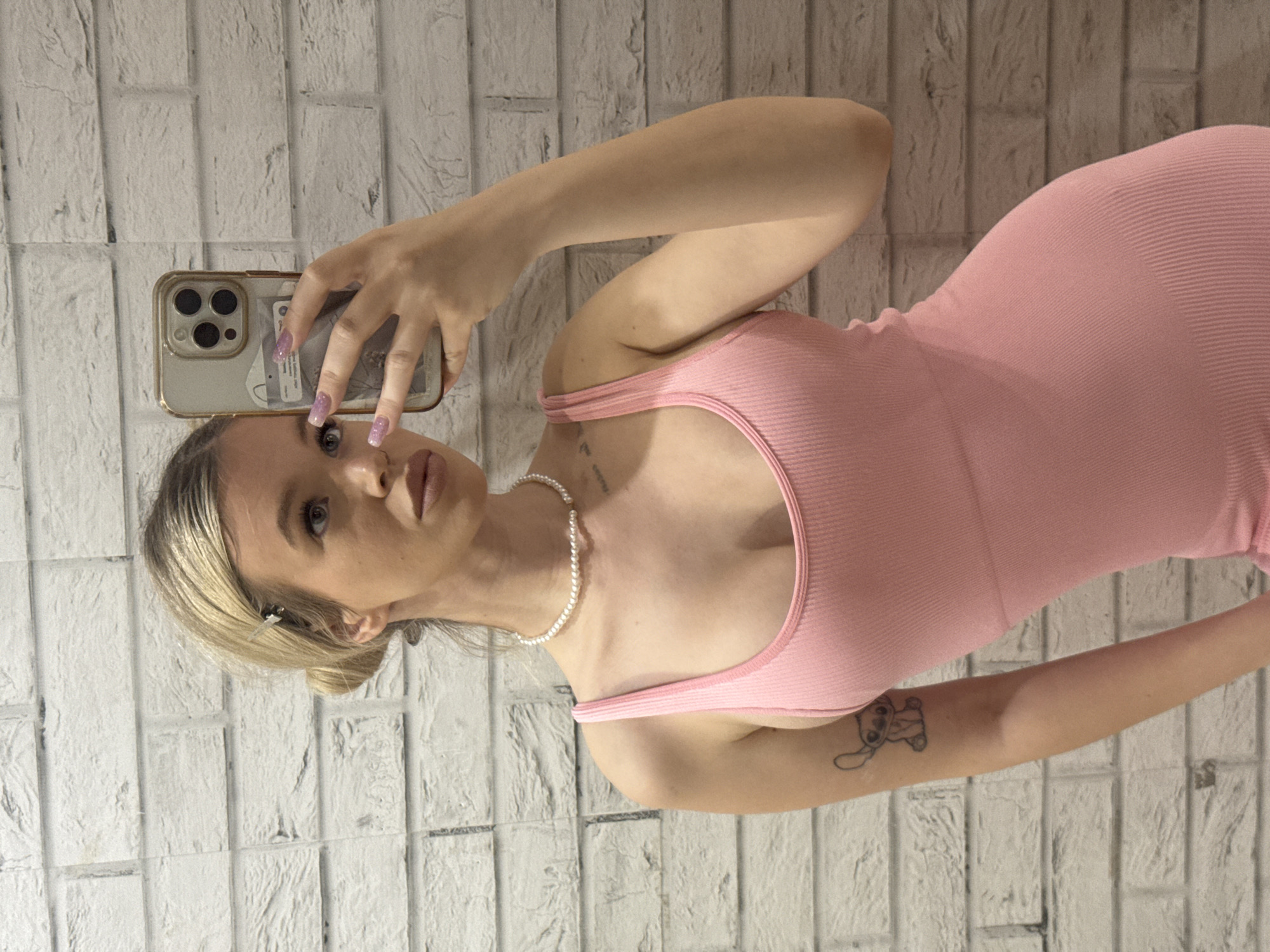 A young woman with blonde hair styled in a bun, holding a phone to take a selfie in a mirror. She is wearing a pink ribbed tank dress and a pearl necklace. She has a Stitch tattoo on her left arm. The background is a white brick wall. The image was taken indoors, likely in a residential setting. There is no information to suggest this location is in Saint Petersburg, Russia.