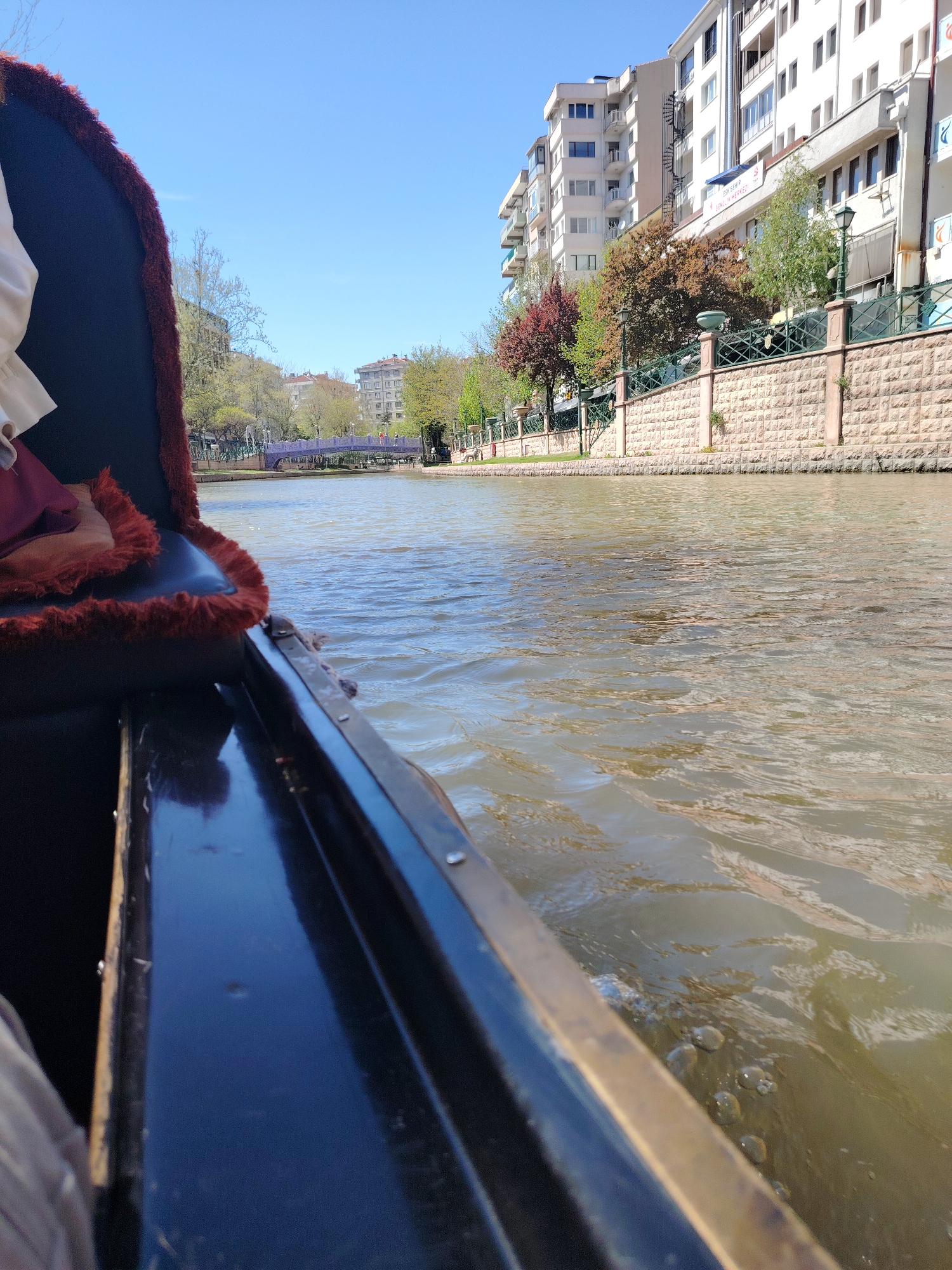 This media file presents a first-person perspective from a boat, resembling a gondola, navigating a canal or river in an urban setting. The boat's interior, visible on the left, features dark blue material with a plush, red fur-like trim. A person's arm and a glimpse of clothing are visible within the boat.
The waterway itself appears greenish-brown with ripples, indicating gentle movement. Along the right bank, a decorative stone-faced wall with a dark metal fence and streetlights separates the water from the land. Behind this wall, trees with a mix of green, red, and brown leaves suggest either spring or early autumn.
Several multi-story residential or commercial buildings, predominantly light-colored, line the bank. On one of these buildings, a white sign with blue text is visible, reading "ESKİŞEHİR GENÇLİK MERKEZİ" (Eskişehir Youth Center) followed by "MA". In the distance, a distinctive purple or lilac-colored arched bridge crosses the canal, with more urban infrastructure beyond.
The sky is clear and bright blue, indicating a sunny day. The scene depicts a calm, recreational activity in an urbanized landscape, specifically in Eskişehir, Turkey, as confirmed by the visible signage.