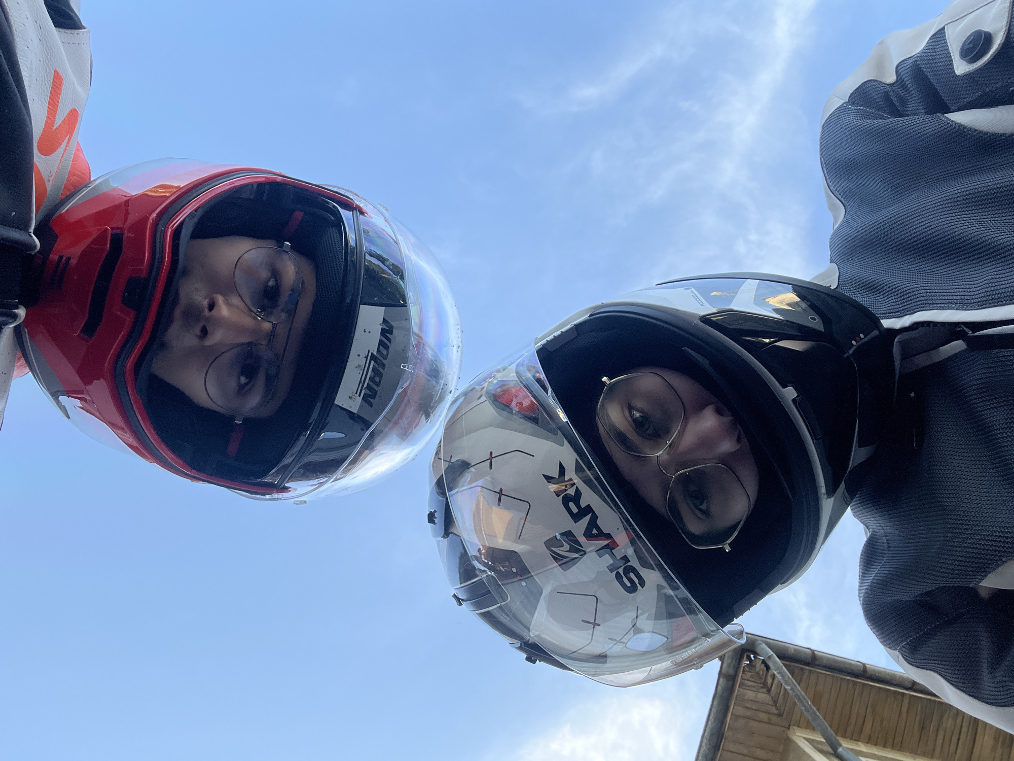 Two people wearing motorcycle helmets are looking down at the camera. The person on the left has a red helmet with the word "NOLAN" visible. The person on the right has a white and black helmet with the word "SHARK" visible. Both people are wearing glasses. The sky is visible in the background, with some clouds. Part of a building is also visible in the lower right corner. The location is Unknown, Unknown.