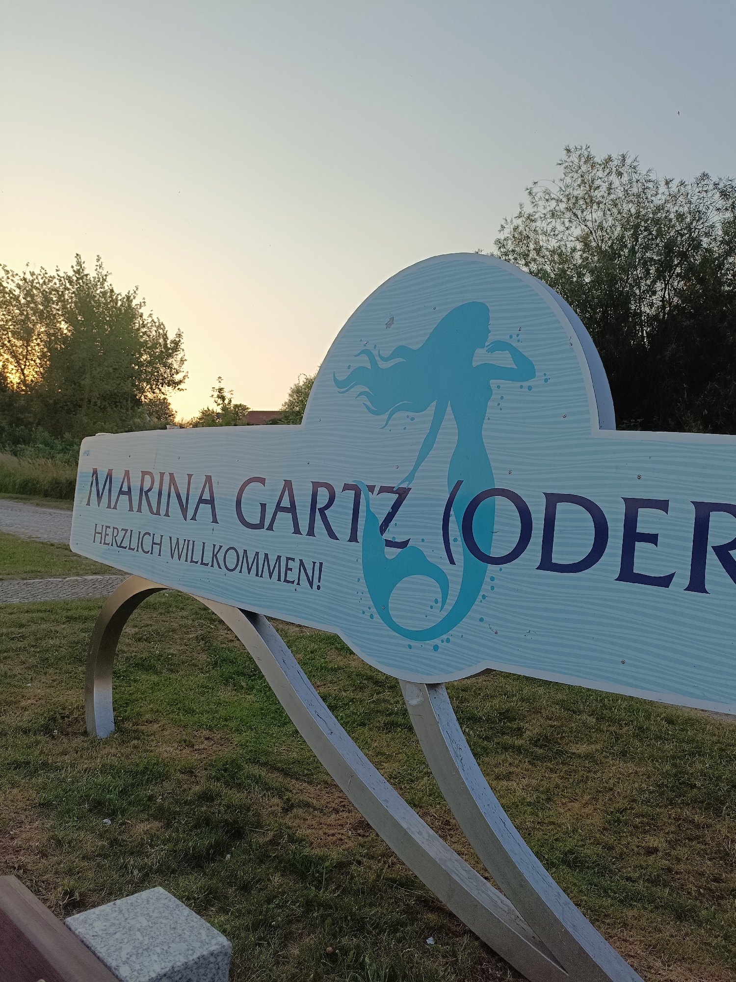 The image shows a light blue sign with a silhouette of a mermaid on it, set against a backdrop of trees and a pale sky, likely at dusk. The sign is mounted on a metal frame and displays text in a dark blue font. The text reads "MARINA GARTZ (ODER)" followed by "HERZLICH WILLKOMMEN!". The setting appears to be an outdoor area with grass and a pathway. The overall impression is that of a welcome sign for the Marina Gartz (Oder) in Germany.
