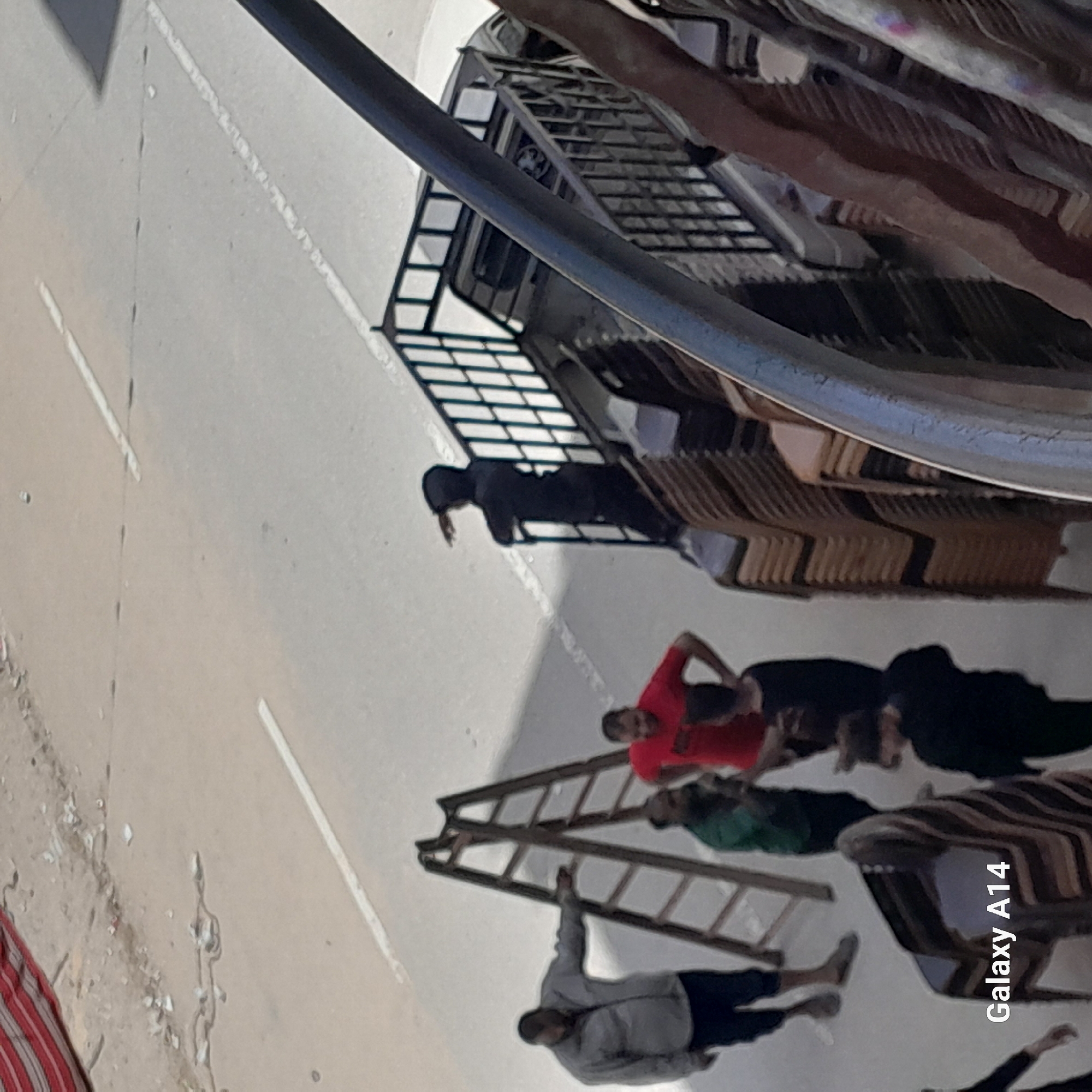 The image is a daytime photograph taken outdoors, presented rotated counter-clockwise. When viewed upright, it depicts an open, light-colored paved area, possibly a street or plaza, with white lines marked on the ground. There are visible strong shadows, indicating direct sunlight and clear weather.
In the mid-ground, on the left, a person dressed in a black hooded garment stands next to a dark metal shelving or display unit. To their right, numerous light-colored chairs are stacked under what appears to be an awning or another part of the metal structure.
Further to the right, a group of three men is visible. One man wears a red t-shirt, another a green t-shirt, and the third a dark shirt. In the foreground, another man wearing a grey hoodie and dark pants is seated or lying on the ground, with his arm extended towards a wooden ladder that lies horizontally on the ground. Some debris or light-colored spatter is visible on the paved surface to the left.
A large, curved metallic structure, possibly a railing or part of a vehicle, dominates the upper right portion of the scene. The overall impression is one of a casual, possibly commercial or public, outdoor space. The text "Galaxy A14" is visible in the bottom right corner, indicating the device used to capture the image. No distinct activities are clearly observable beyond people standing or resting.
