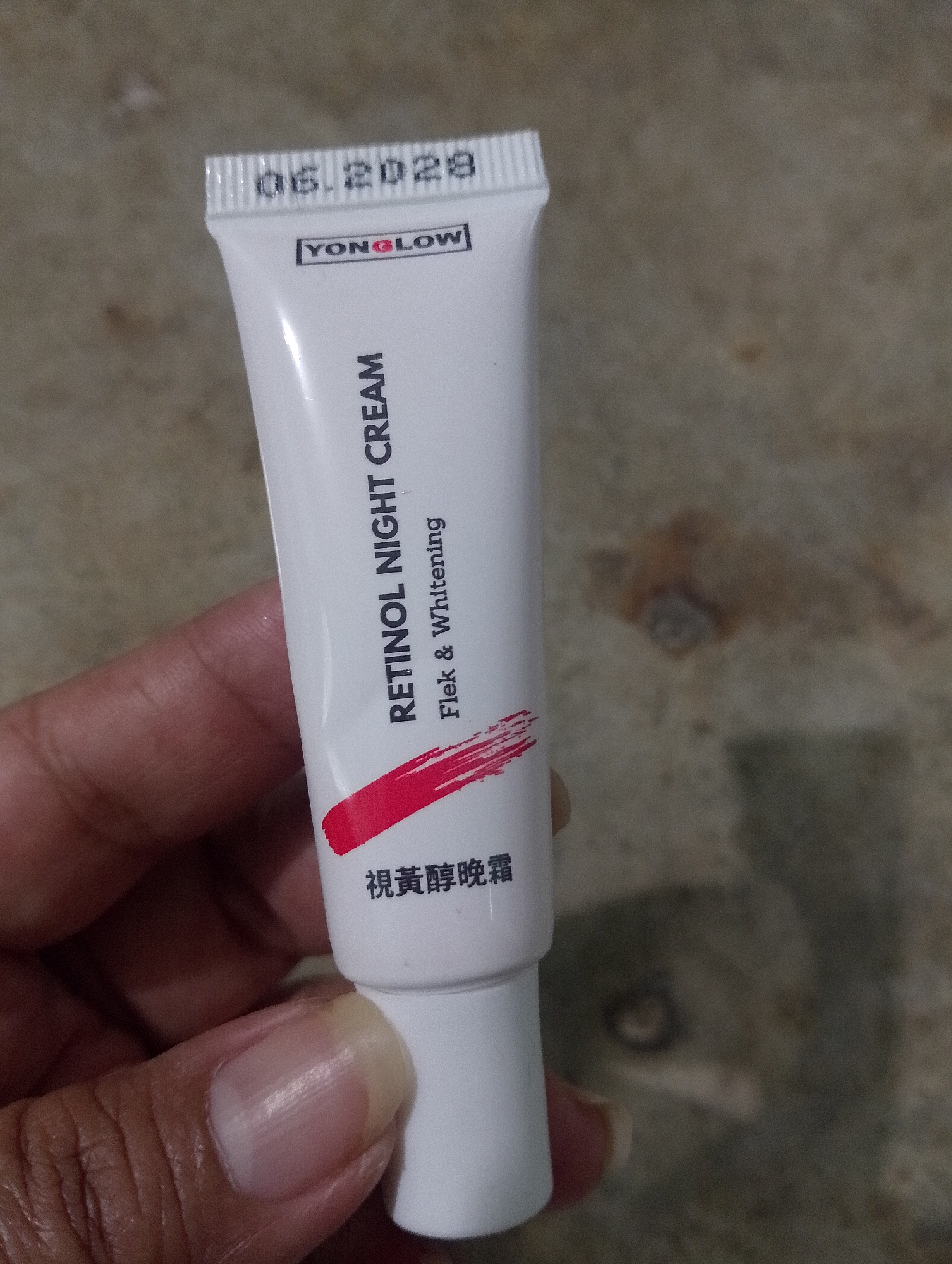 An expired tube of YONGGLOW Retinol Night Cream with Flek & Whitening is being held by a person's hand. The expiration date on the tube reads "06.2029". The background is a textured surface that appears to be a floor or wall. There is no indication of the location being in the city of Serbelawan, Indonesia.