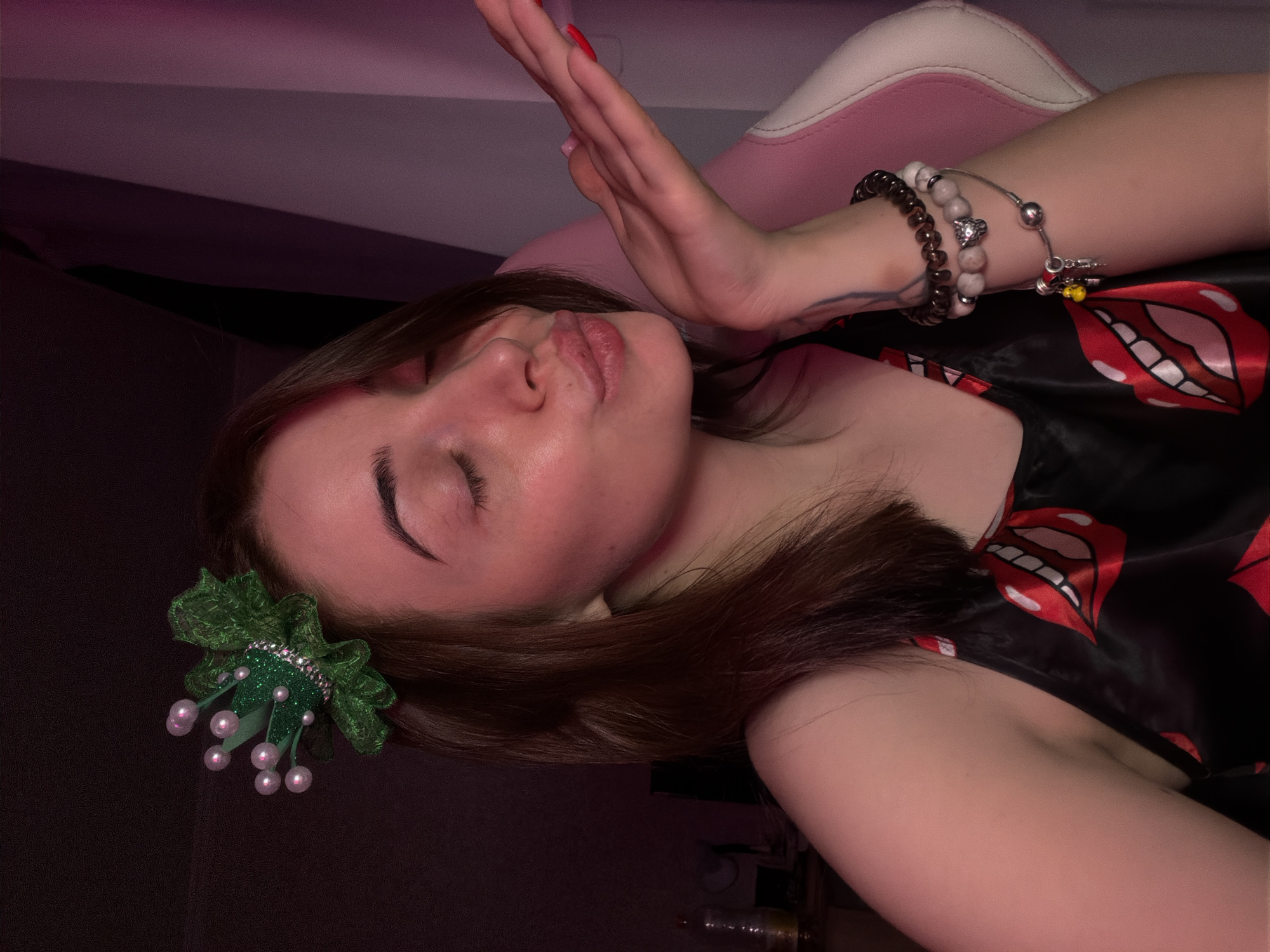 A young woman with medium-length brown hair is shown from the chest up, her eyes closed, blowing a kiss towards the camera. She is wearing a black satin top with a pattern of red and white lips. On her head, she wears a green sparkly hair accessory resembling a crown with pearl-like decorations. Her left hand is extended, palm open, towards the camera. She has several bracelets on her right wrist, including one with a lion's head charm. The background is dark, with a pink hue emanating from the right side, suggesting ambient lighting. She is seated in a pink and white chair. The image quality is low, with visible noise and artifacts. The location is not specified.