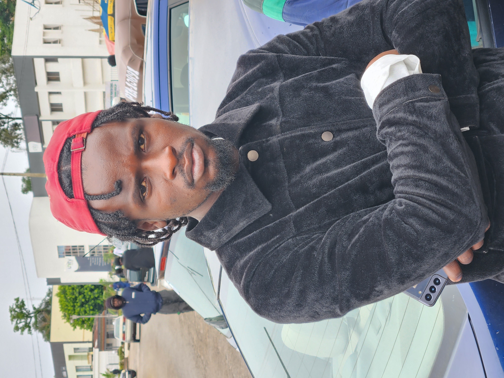 A Black man with dark skin, dreadlocks, and a beard is the central figure, looking directly at the camera with a neutral expression. He wears a red baseball cap backward, a dark, plush-looking jacket (possibly black or dark grey), and a white shirt whose cuffs are visible. His arms are crossed in front of him. A smartphone with multiple camera lenses is partially visible, tucked into his left jacket pocket.
The man is positioned outdoors in what appears to be an urban or suburban street in Jos, Nigeria, leaning against the rear of a blue sedan. In the background, another Black man in a dark blue top and dark trousers stands near a white car. Further back, various buildings are visible, including a prominent white structure with several windows and a light-colored building. Trees and other foliage are scattered throughout the scene.
The image captures the man posing for a photograph during daytime, under what appears to be overcast conditions, resulting in soft, diffused lighting. The overall mood is casual. Blurry, indistinct white text is visible on a brown banner-like structure behind the blue car, and another blurry sign on the white building in the mid-ground contains unreadable text, possibly including a logo or company name.