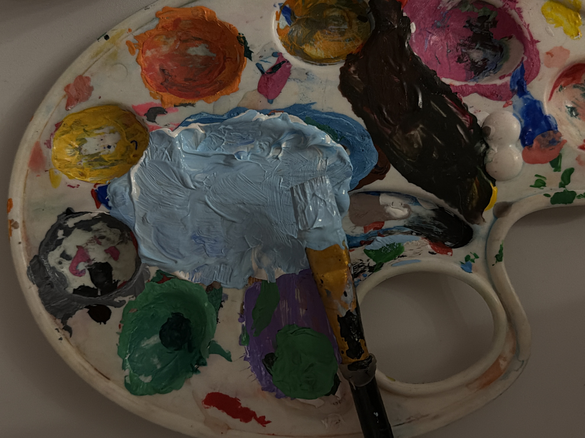 A painter's palette sits on a white surface, covered in a variety of colors of wet paint. From top left, clockwise: orange, yellow, gray, green, red, purple/green mixture, light blue. A paint brush is laid across the palette, handle pointing toward the bottom of the frame, with light blue paint on the bristles. The palette is white and plastic, with circular wells for holding paint. The background appears to be a plain white wall. There is no indication of the location being Perm, Russia.