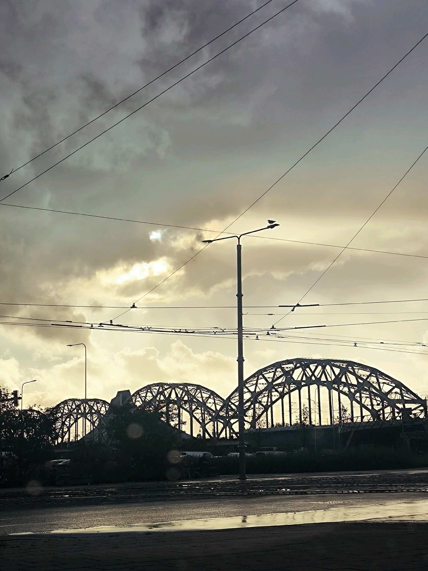 This is a photograph of a bridge in Riga, Latvia, taken at dusk. The sky is filled with dramatic, cloudy textures, with the sun breaking through in a few spots, creating a soft, golden light. The bridge itself is a large, arched structure made of steel, with intricate latticework. Silhouetted against the sky, the bridge appears imposing and grand.
In the foreground, there are streetlights and overhead wires, suggesting an urban environment. The road in front of the bridge is wet, reflecting the light and the overcast sky, indicating recent rain or damp conditions. Trees and darker structures are visible behind the bridge, adding depth to the scene. The overall mood of the image is atmospheric and somewhat melancholic, typical of a moody twilight in a European city. There are no people clearly visible, but the presence of infrastructure like streetlights and wires implies human activity and a lived-in urban setting. The scene is quiet and still, capturing a moment of transition between day and night.