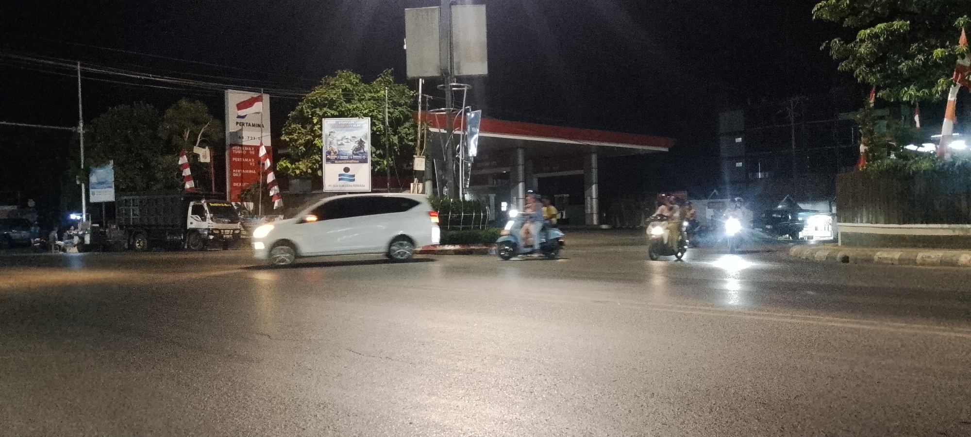 The image depicts a nighttime street scene at an intersection in Batulicin, Indonesia. A white minivan is traveling across the intersection from left to right, with its headlights illuminated. To the left of the minivan, a dark-colored truck is parked. Several people are riding motorcycles, some with passengers, moving through the intersection. One motorcycle, in the foreground on the right, has its headlight brightly lit, illuminating the wet pavement. In the background, a Pertamina gas station canopy is visible, with red and white signage and flags. Trees and a building are also present in the background. The road surface appears wet, reflecting the light from the vehicles and streetlights.