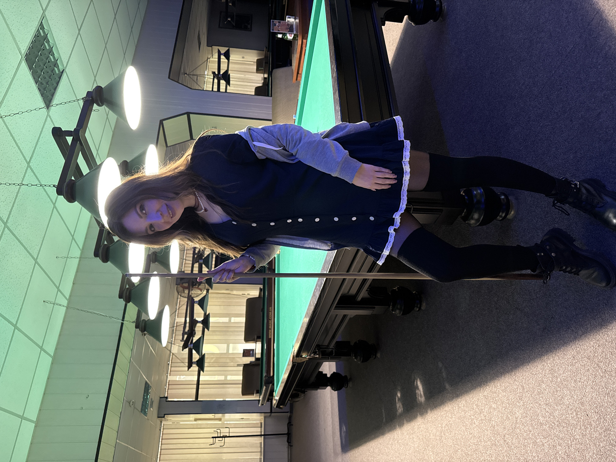 A young woman is posing next to a billiards table in what appears to be a billiards hall. She has long brown hair, is wearing a dark dress with a white trim, a gray hooded sweatshirt, black thigh-high socks, and black boots. She is holding a pool cue and smiling at the camera. The billiards table has a green felt surface and is surrounded by black trim and legs. Above the table hang a series of lamps with green shades. The walls of the hall are a light green color with a diamond pattern. The floor is carpeted in gray. Through a window, rows of chairs and tables can be seen in the background. This scene is in Kryvyi Rih, Ukraine.