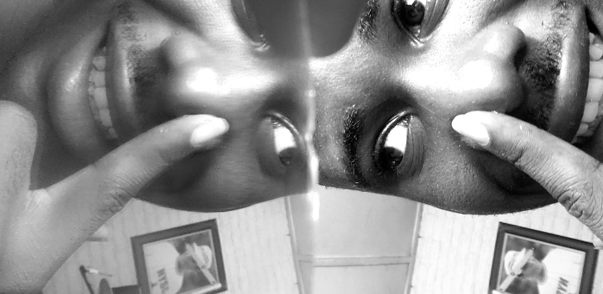The black and white photograph depicts a distorted, close-up view of a person's face, duplicated through reflection, seemingly taken from an unusual perspective such as lying down. The individual has dark skin and is smiling widely, exposing their teeth. A finger is pointing towards the eye of each reflected face. In the background, visible below the faces, are framed pictures on a wall and a bathroom-like setting with tiled walls. There is no explicit evidence to place this photo in Narayi, Nigeria, only the description of the image.