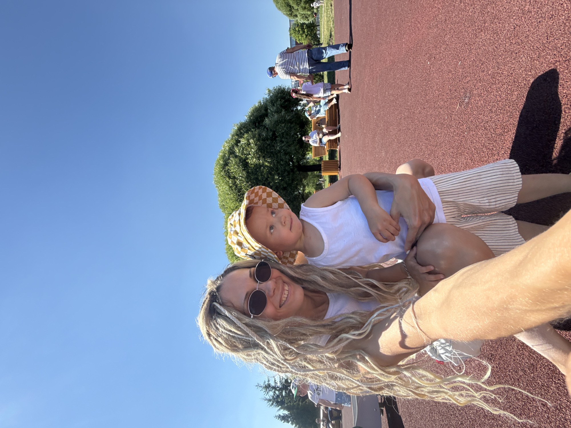 In an outdoor park setting in Zavolzhye, Russia, a woman with blonde dreadlocks and sunglasses smiles at the camera while holding a young child. The child, wearing a checkered hat and a white tank top, looks away from the camera. The woman is wearing a white tank top. In the background, other people are visible: a man in a striped shirt and blue jeans walks with a young girl in a pink headband and white shirt, while another woman and child sit on a bench under a large tree. The ground of the park is a red, textured surface, and the sky is clear blue.