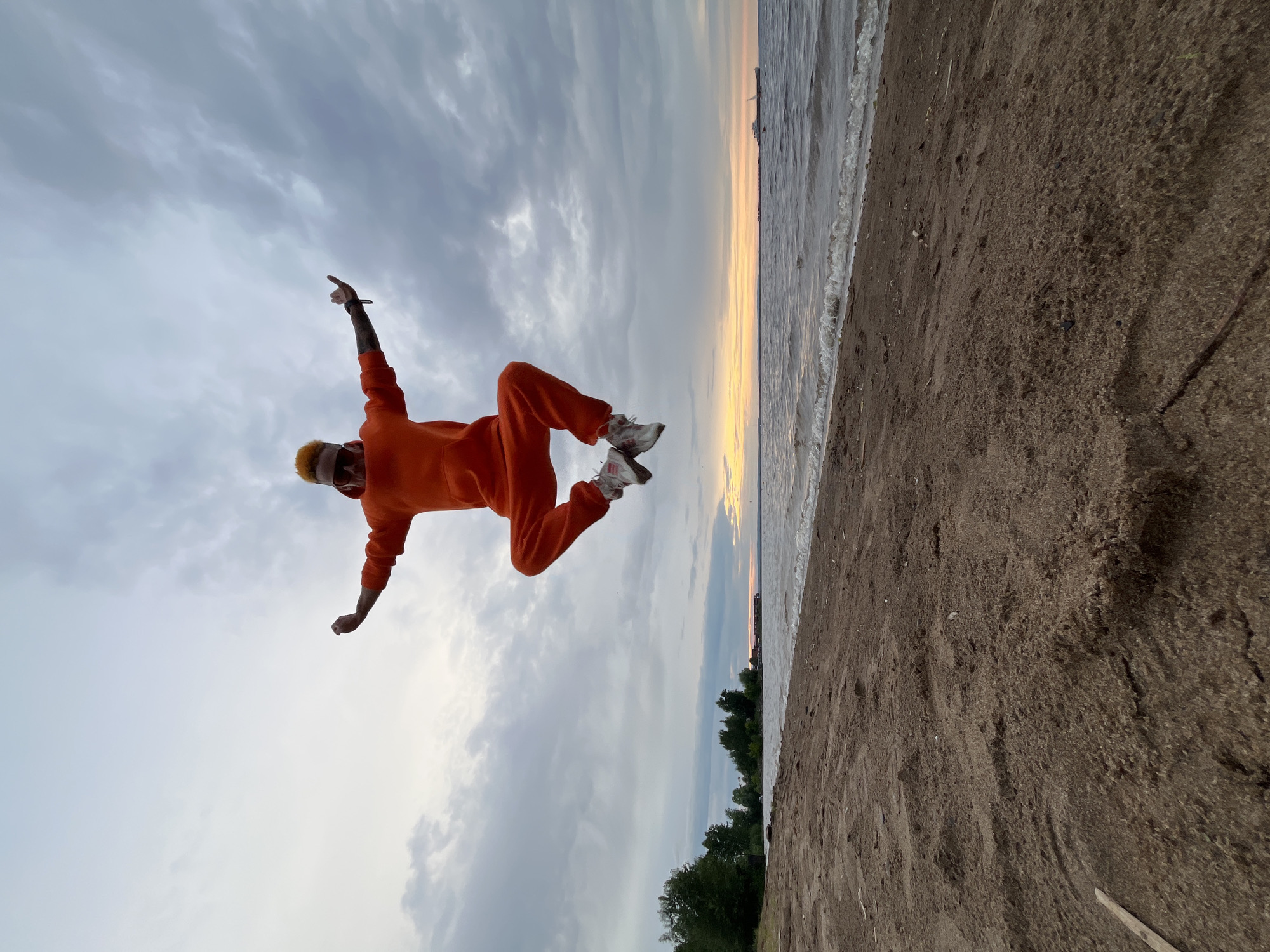 The image shows a person in mid-air, performing a jump on a sandy beach beside a body of water. The person is wearing an orange jumpsuit and white sneakers, with bright orange hair. Their arms are outstretched, with one pointing upwards and the other bent. The setting is a beach with dark sand and gentle waves lapping at the shore. In the distance, a line of trees separates the beach from the sky. The sky is filled with dramatic, cloudy formations, with a hint of sunset colors near the horizon. The overall impression is one of dynamism and freedom, with the person appearing to defy gravity. The time of day appears to be late afternoon or early evening, given the soft light and the colors in the sky. There is no visible text in the image. The location is Chaikovsky, Russia.
