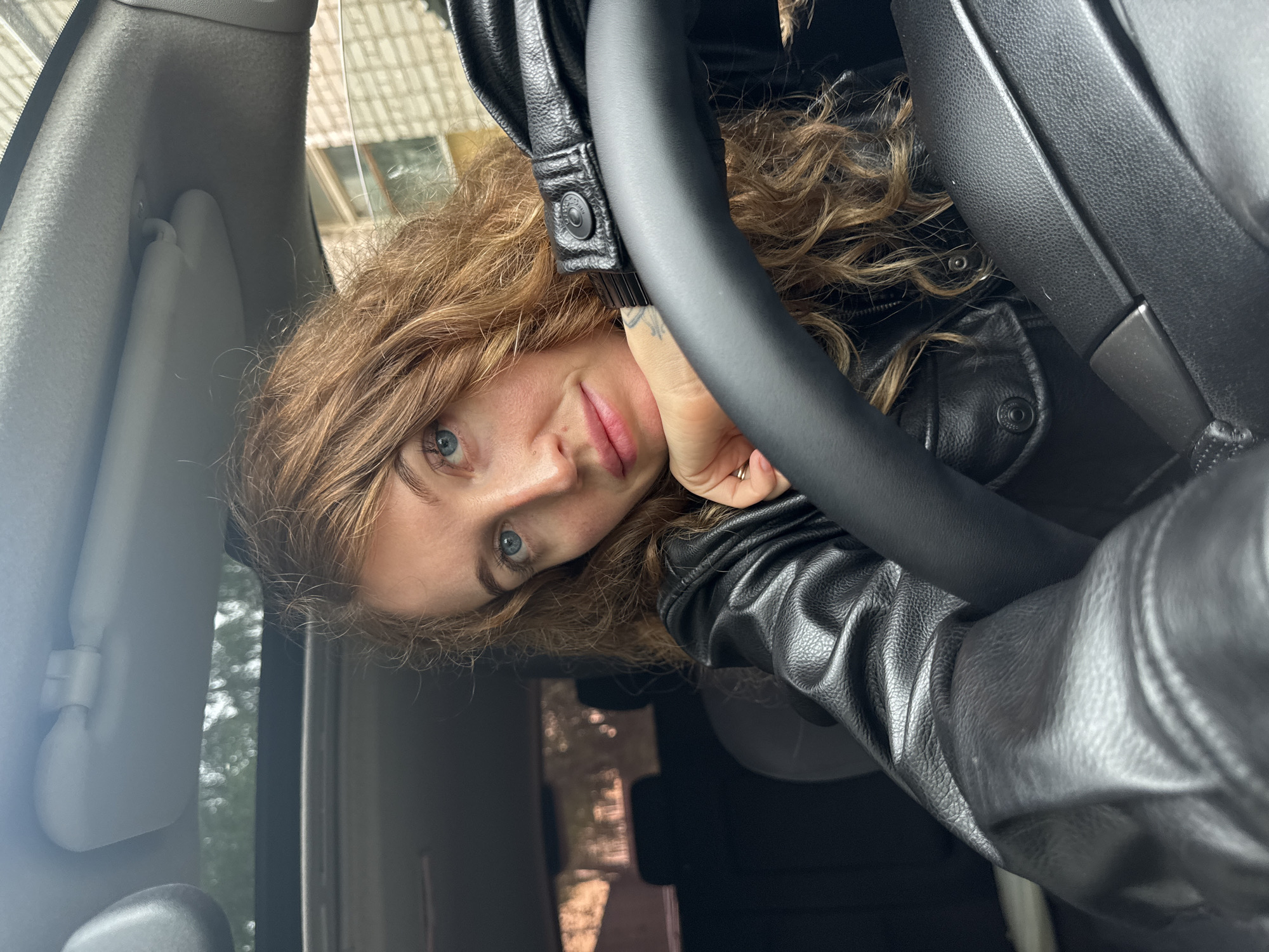 The image, taken in the city of Chaikovsky, Russia, displays a female subject positioned inside the driver's seat of a vehicle. The woman has long, wavy, reddish-brown hair and light blue eyes. She is wearing a black leather jacket, with a prominent snap button visible on the sleeve cuff of her right arm. Her left hand, adorned with a silver-toned ring on the ring finger, supports her chin, while her right forearm and wrist rest on the top segment of a black steering wheel. A dark-colored band is visible on her right wrist, and a faint tattoo is partially visible on her right forearm below the band. The car's interior features a dark-colored dashboard and seating. Overhead, a sunroof is visible, through which blurred green foliage can be seen. To the right, a grey sun visor is present, and through the passenger side window, a light-colored brick or stone building facade is partially visible. The subject is looking directly towards the viewer.