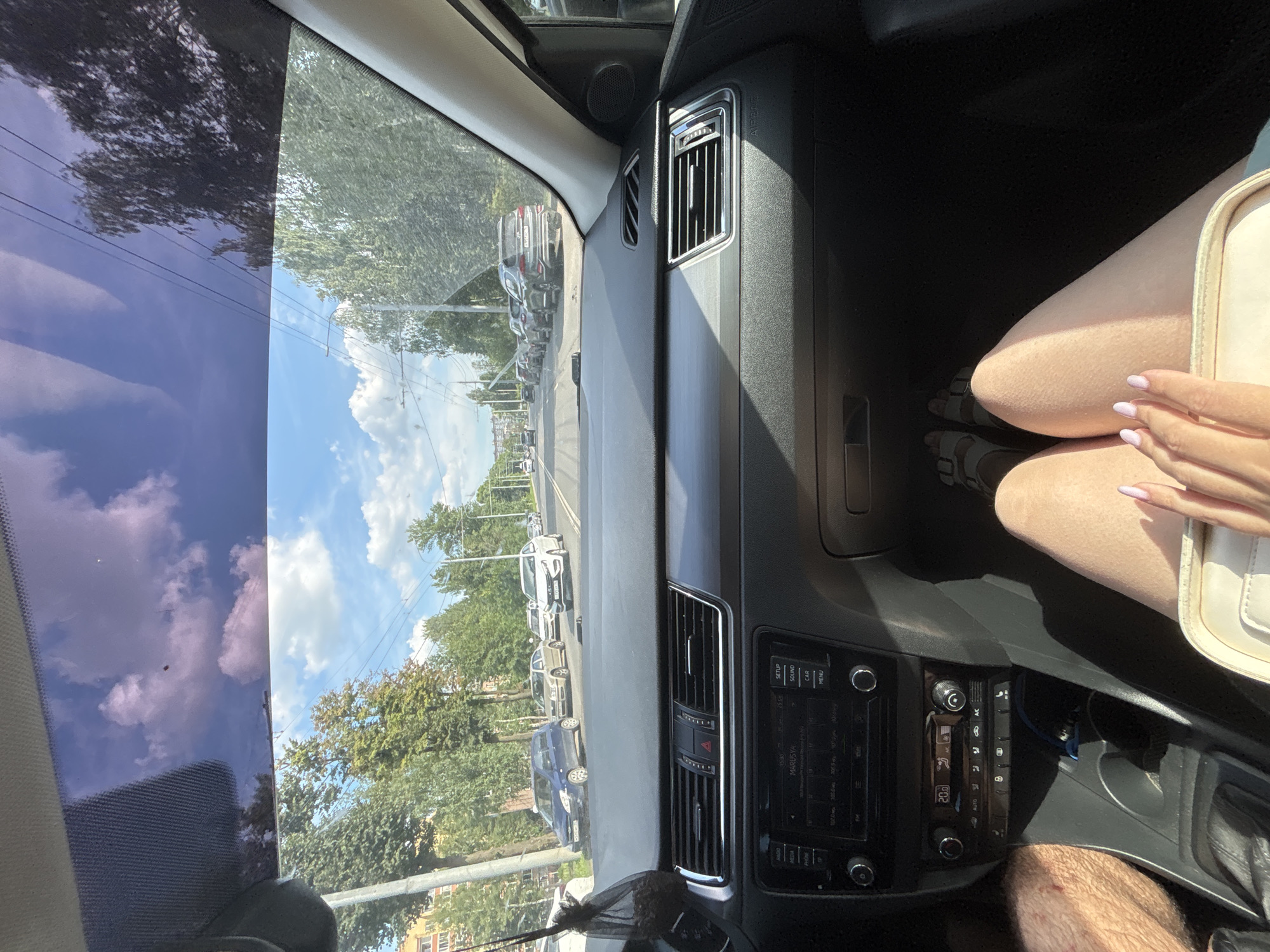 The image shows the interior of a car, looking forward through the windshield and side windows. The view outside the car reveals a sunny day in a city, with cars on a road lined with trees and buildings. In the foreground, the dashboard of the car is visible, including the radio and air conditioning controls. A person's legs and feet are visible in the passenger seat, and their hand rests on a light-colored bag. The location is identified as Rusanovo, Russia.