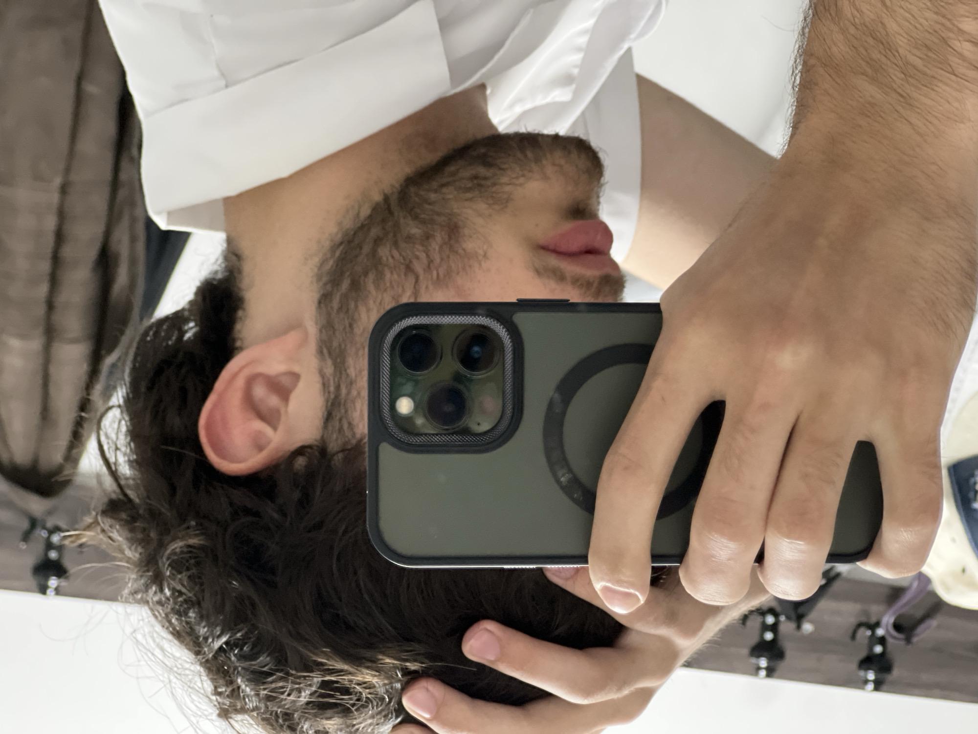 A man is taking a selfie in a mirror. He is wearing a white collared shirt. He has dark hair and a beard. He is holding a dark green iPhone with a MagSafe ring on the back in his right hand. His left hand is visible resting on his head. The reflection shows a coat rack with coats and a white surface that reflects some of the objects in the room. There is no indication of this photo being taken in Yerevan, Armenia.