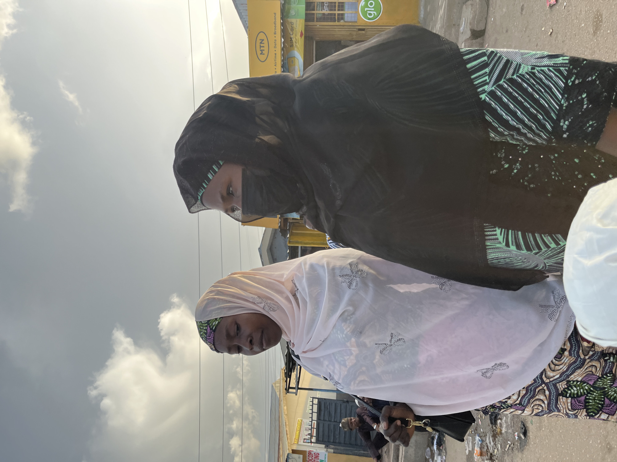 An outdoor street scene in Gura Top, Nigeria, is captured under a partially cloudy sky, with soft natural light suggesting early morning or late afternoon. Two adult women, both of dark complexion, stand in the foreground, facing slightly towards the right.
The woman on the left wears a colorful patterned head covering and a light pink translucent shawl adorned with silver, crystalline embellishments over a patterned dress. She holds a small black handbag with a gold-colored clasp or keychain in her visible left hand and looks downwards with a somber expression.
The woman on the right wears a black head covering (hijab) and a black face mask, with her eyes looking towards the right. Beneath her black outer garment, a black and green patterned fabric is visible.
In the left background, a dark-skinned male figure with a light-colored cap is seated on the ground, leaning against a building with a dark metal gate. A partially visible white sign with green and black text is affixed to the building.
The right background features prominent commercial signage, including a bright yellow "MTN" sign displaying "o Airtime • Data • Broadband" and "SIM Card" text, and below it, a yellow sign with a green "glo" logo. These signs indicate mobile telecommunication service providers. Simple building structures are visible behind these signs.
The ground in the foreground and mid-ground is uneven, appearing to be dirt or rough pavement, and is heavily littered with plastic bottles, papers, and other refuse. Thin, dark utility wires are visible horizontally across the upper portion of the sky, which displays a large white cloud formation on the left.