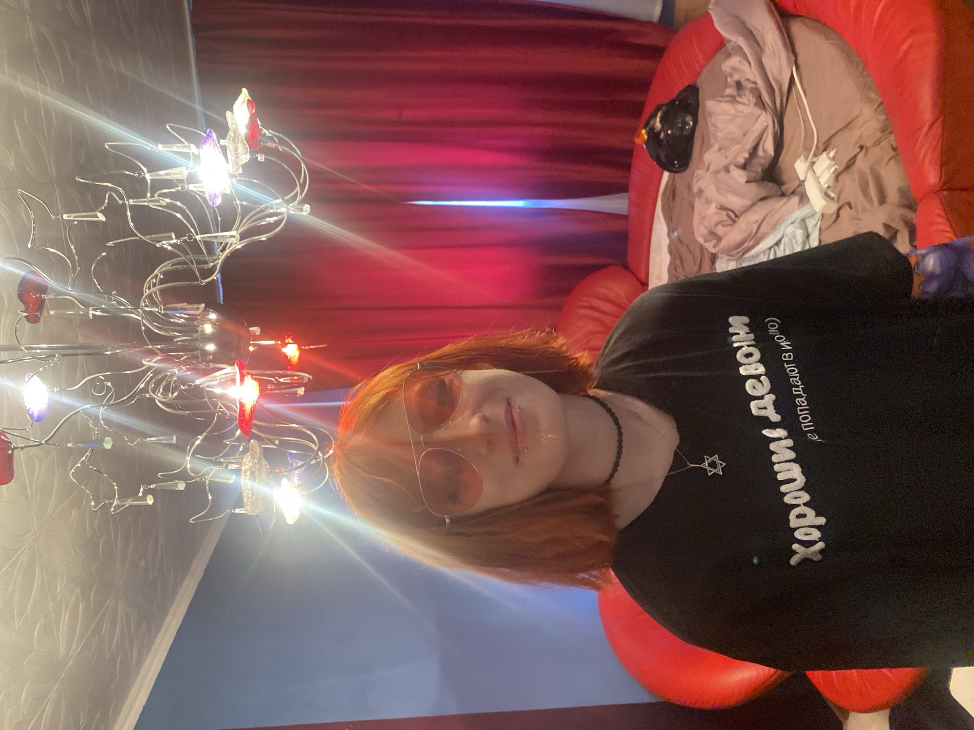 A young woman with short, reddish-orange hair and pink aviator sunglasses stands in what appears to be a bedroom. She is wearing a black t-shirt with white text in Russian, a black choker necklace, and a Star of David pendant. She has a piercing on her lower lip. She is standing in front of a large, circular red couch or bed. Behind her, red curtains are drawn. A distinctive, ornate chandelier with red and purple elements hangs from the textured ceiling. The overall impression is of a dimly lit, possibly theatrical or bohemian, personal space. There is no information that allows for the identification of the city of Tver, Russia.