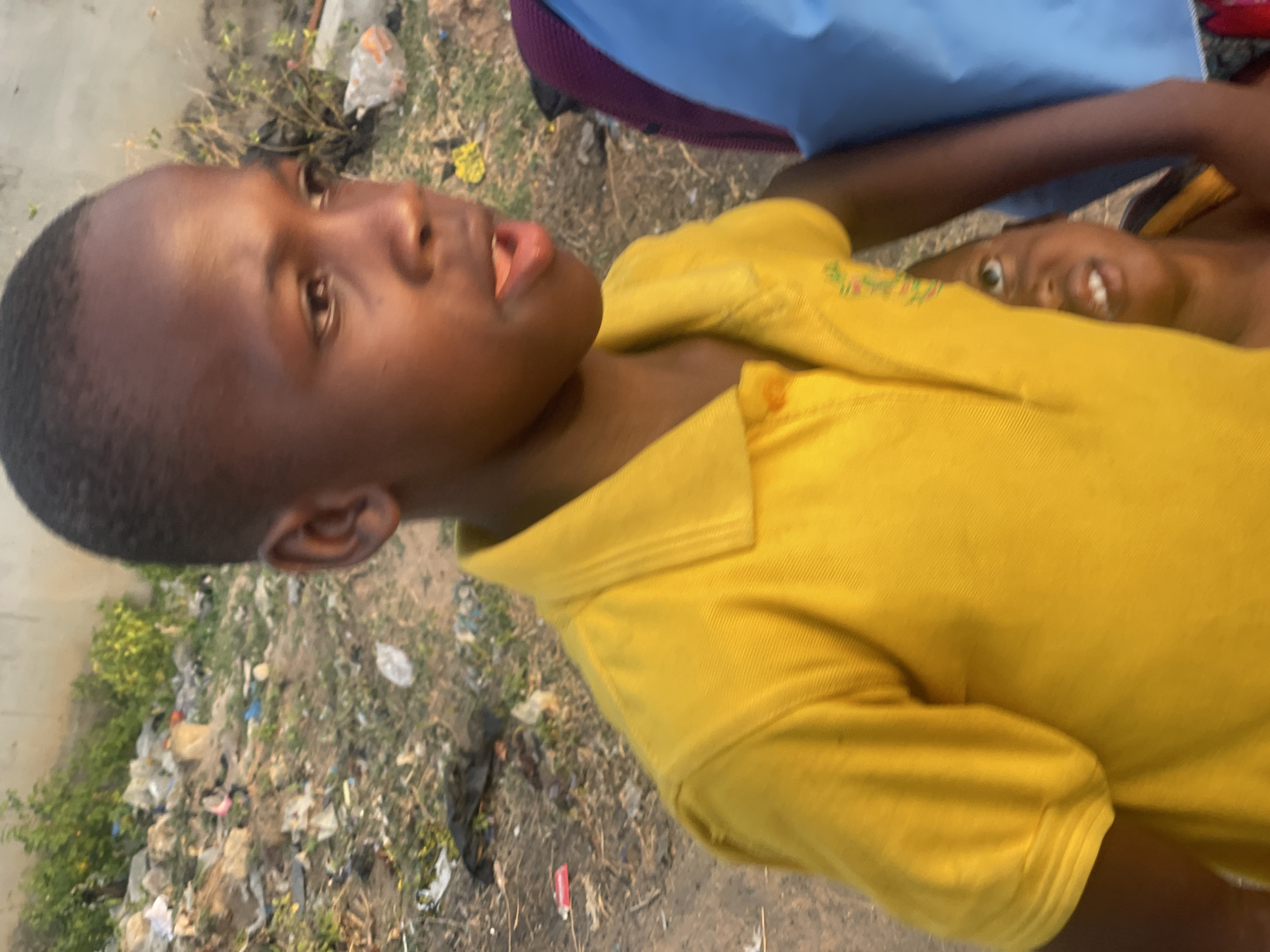 A young boy with dark skin and short hair wearing a yellow collared shirt is looking to the right. Another child is partially visible behind him. The ground appears to be littered with trash and vegetation. The image was taken in daylight. There is no explicit information indicating that the location is Lokoja, Nigeria.